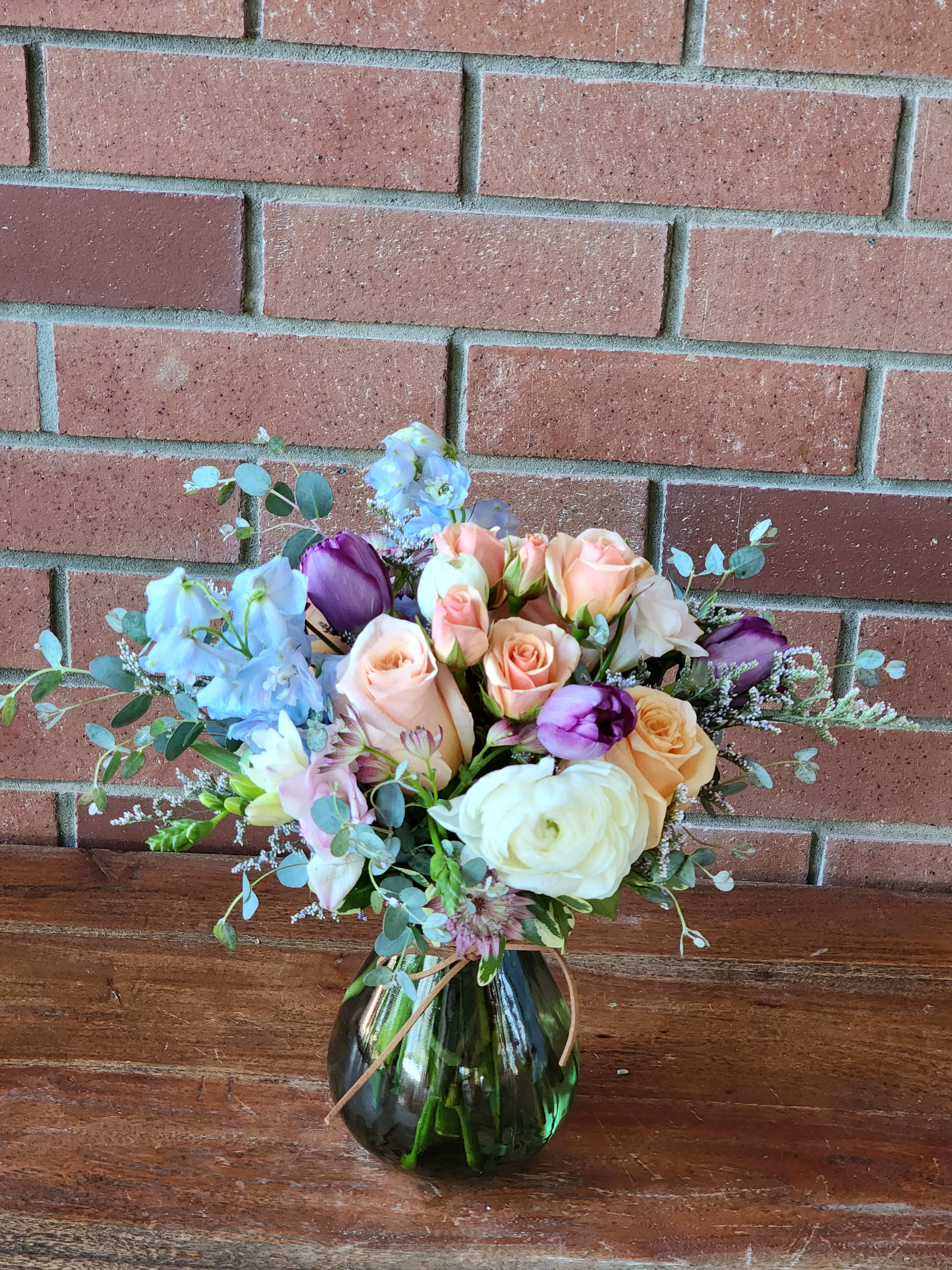 Peach roses, white ranunculus, and purple tulips arranged in a round green glass vase.