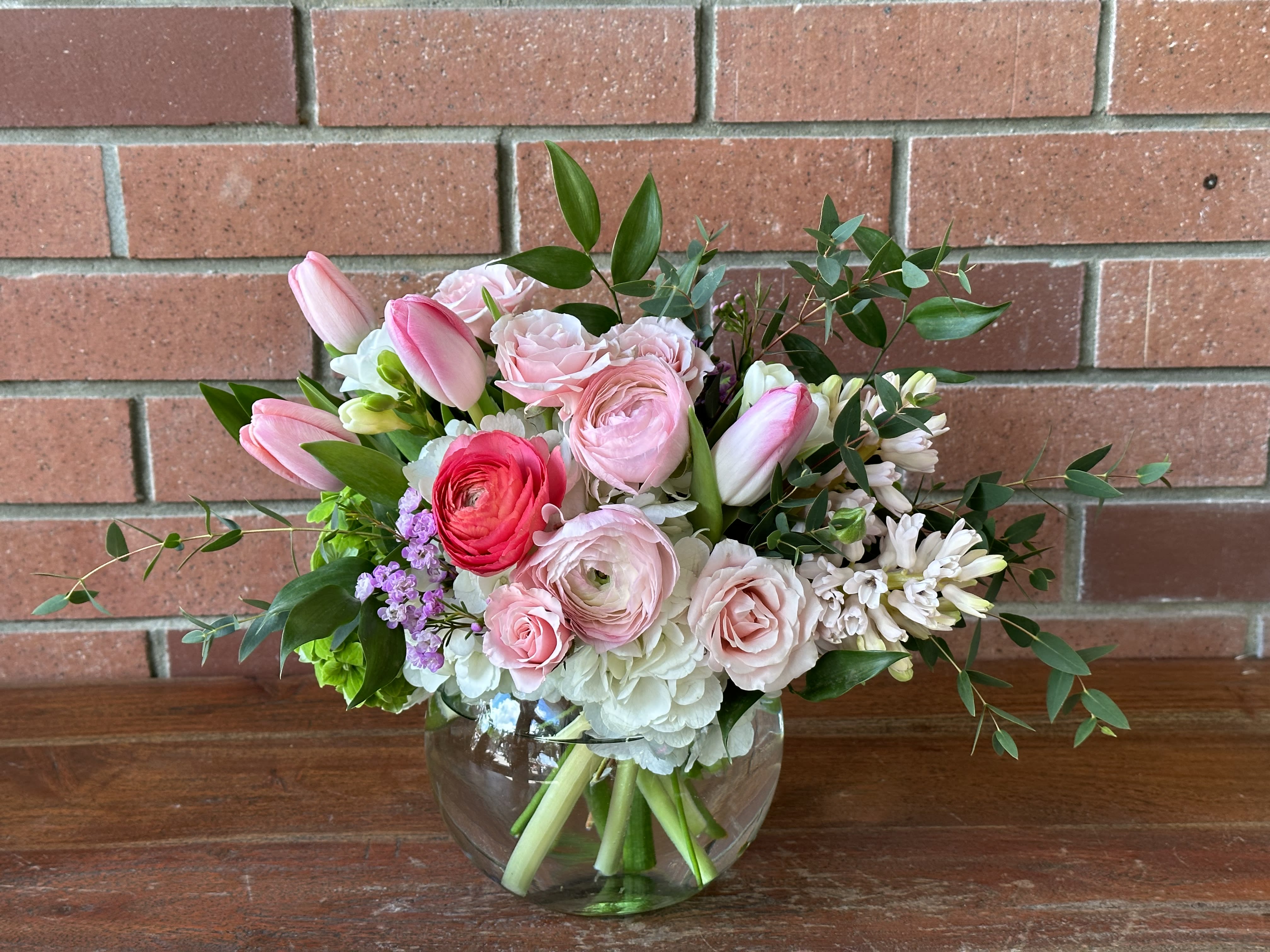 Low glass vase of pink tulips, ranunculus, roses, and white blooms in a rounded bouquet.
