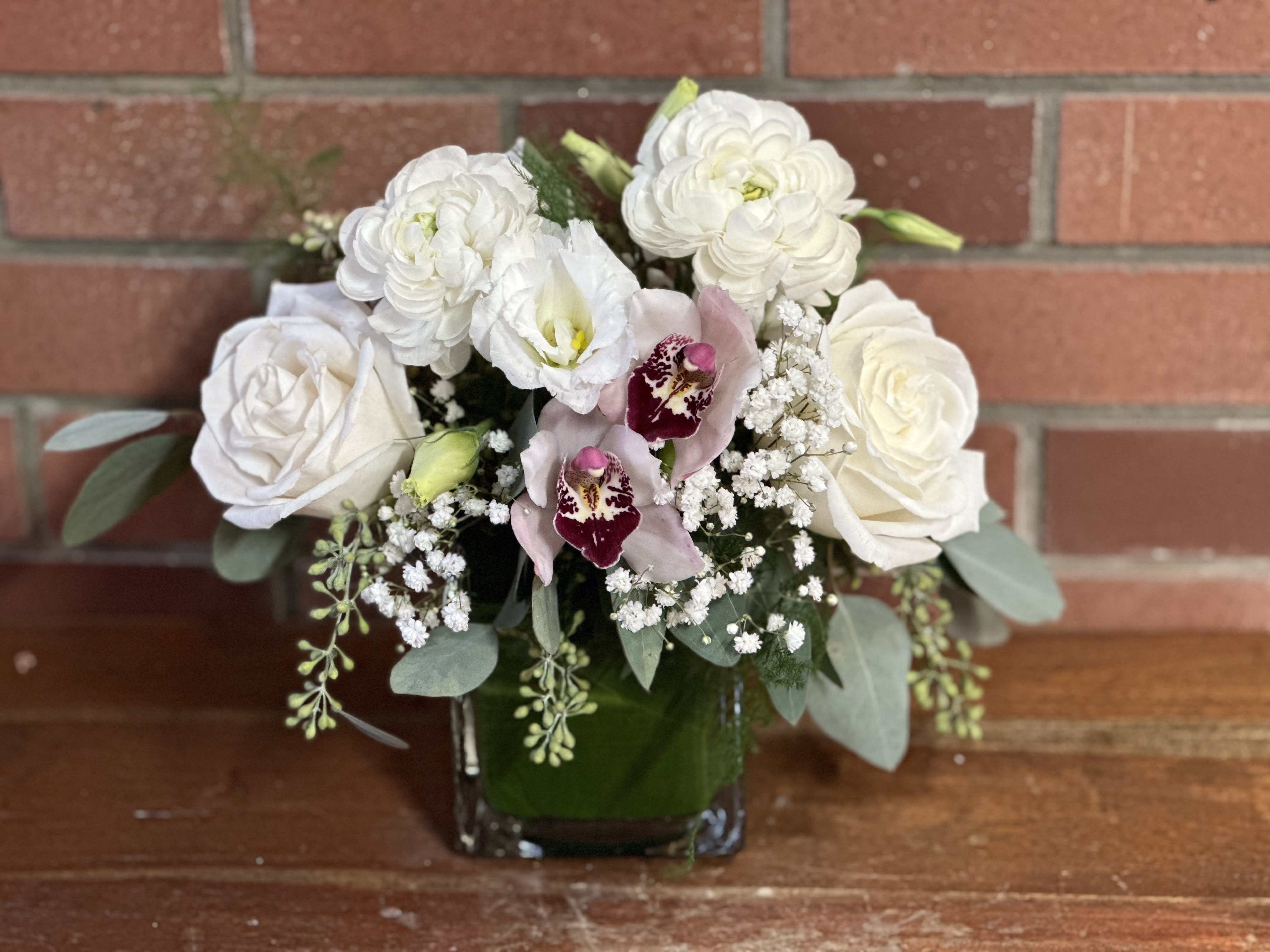 Compact arrangement of white roses and ranunculus with pink orchids in a square glass vase
