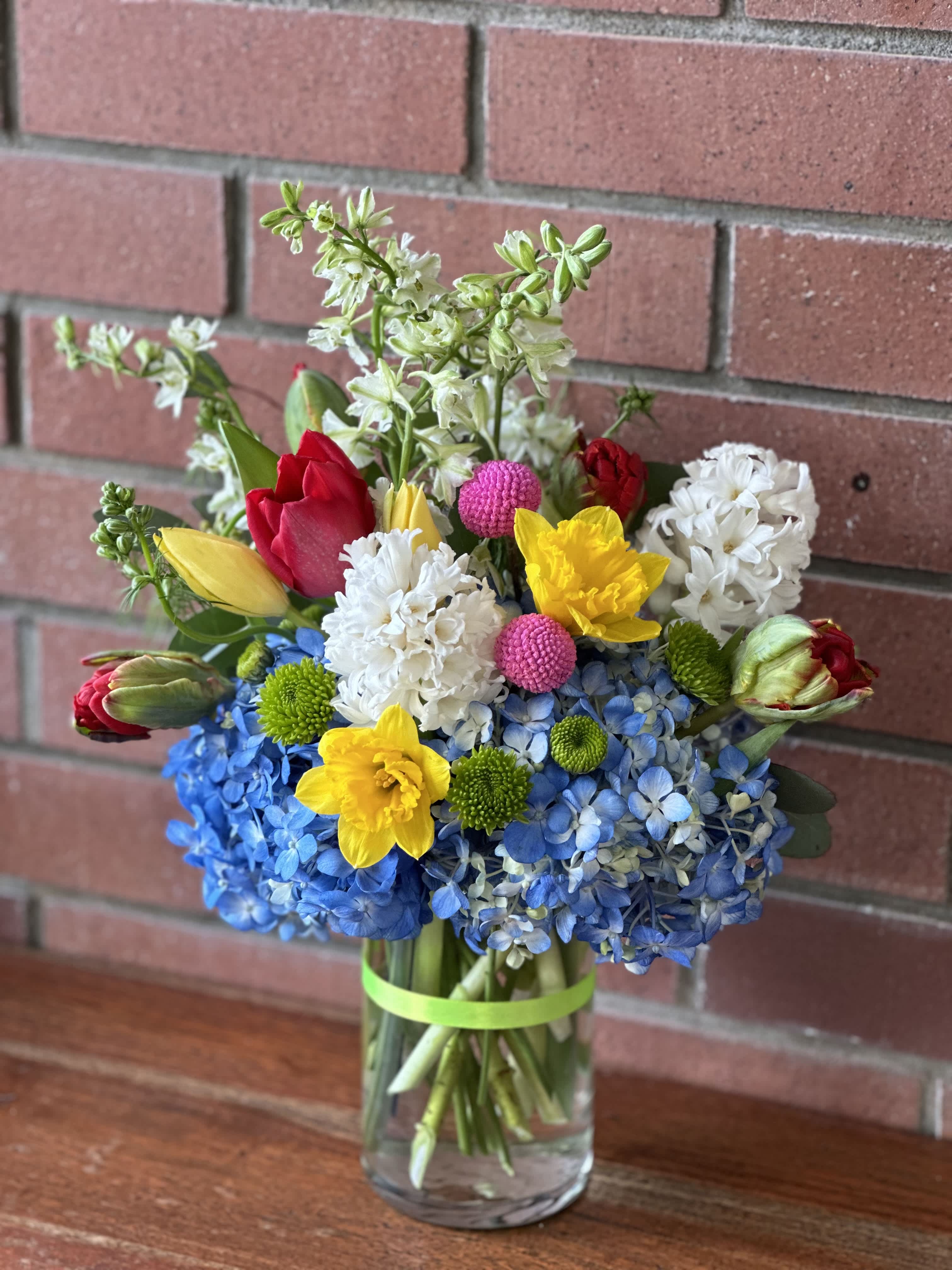 Bright mixed bouquet with blue hydrangeas, yellow daffodils, tulips, and white flowers in a clear glass vase