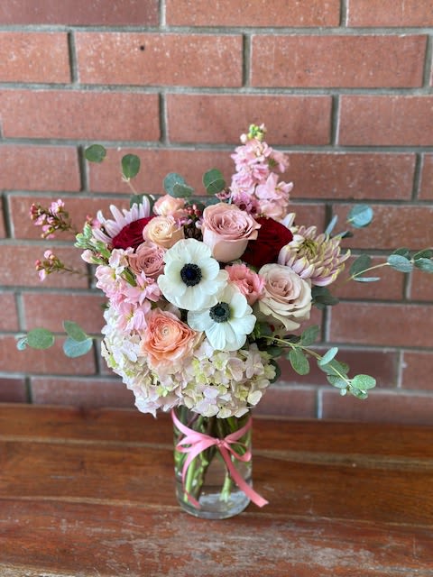 Tall mixed bouquet of soft pink, white, and peach flowers in a clear vase with a pink ribbon.