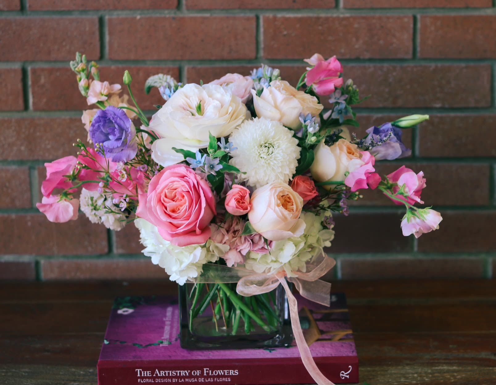 Low pastel arrangement of pink roses and mixed blooms in a square glass vase with ribbon.