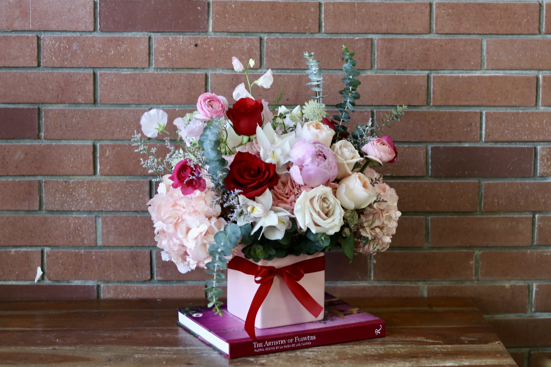 Mixed bouquet of roses, peonies, and hydrangeas in a pink box with a red ribbon