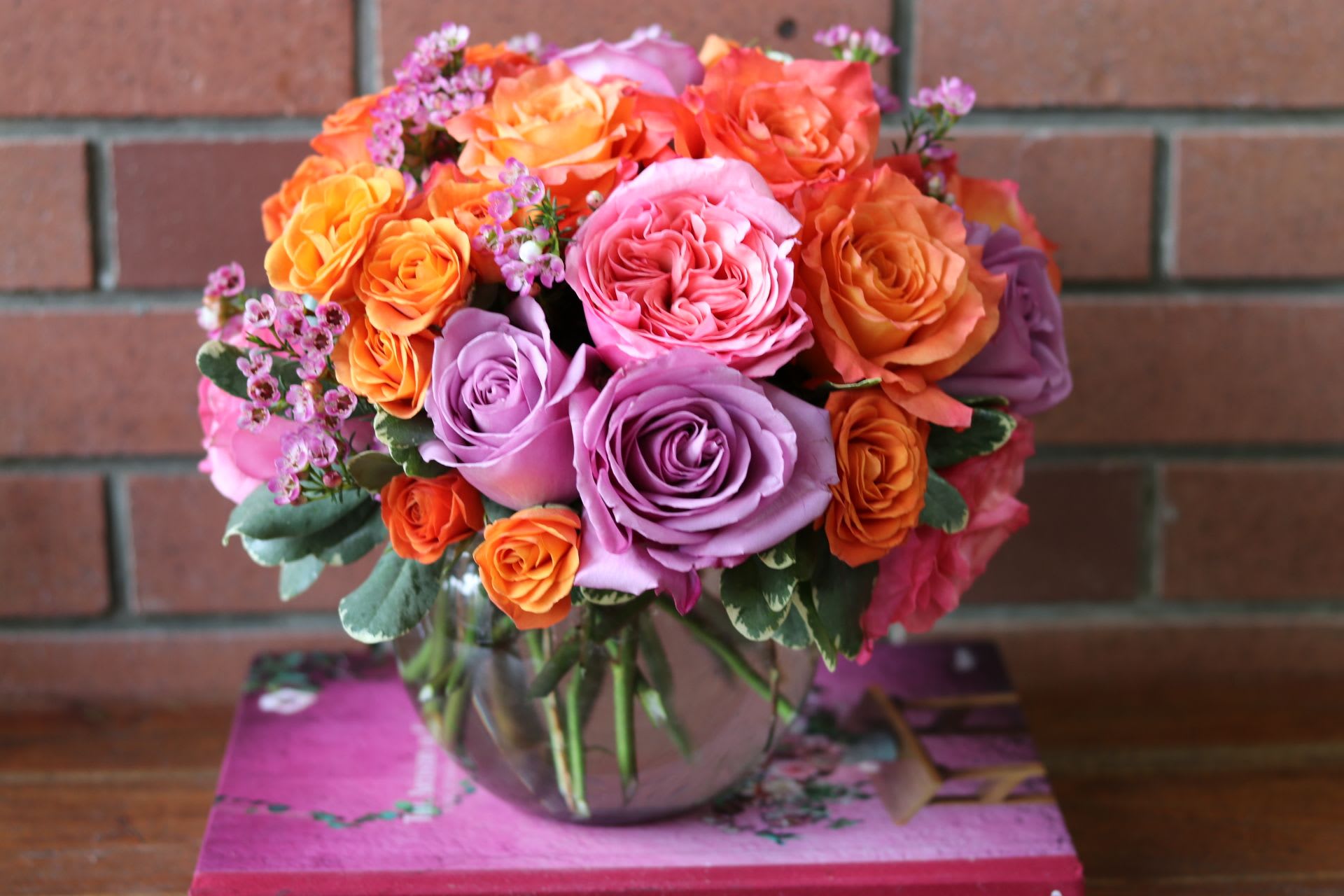 Round arrangement of orange, pink, and lavender roses in a clear glass bowl vase