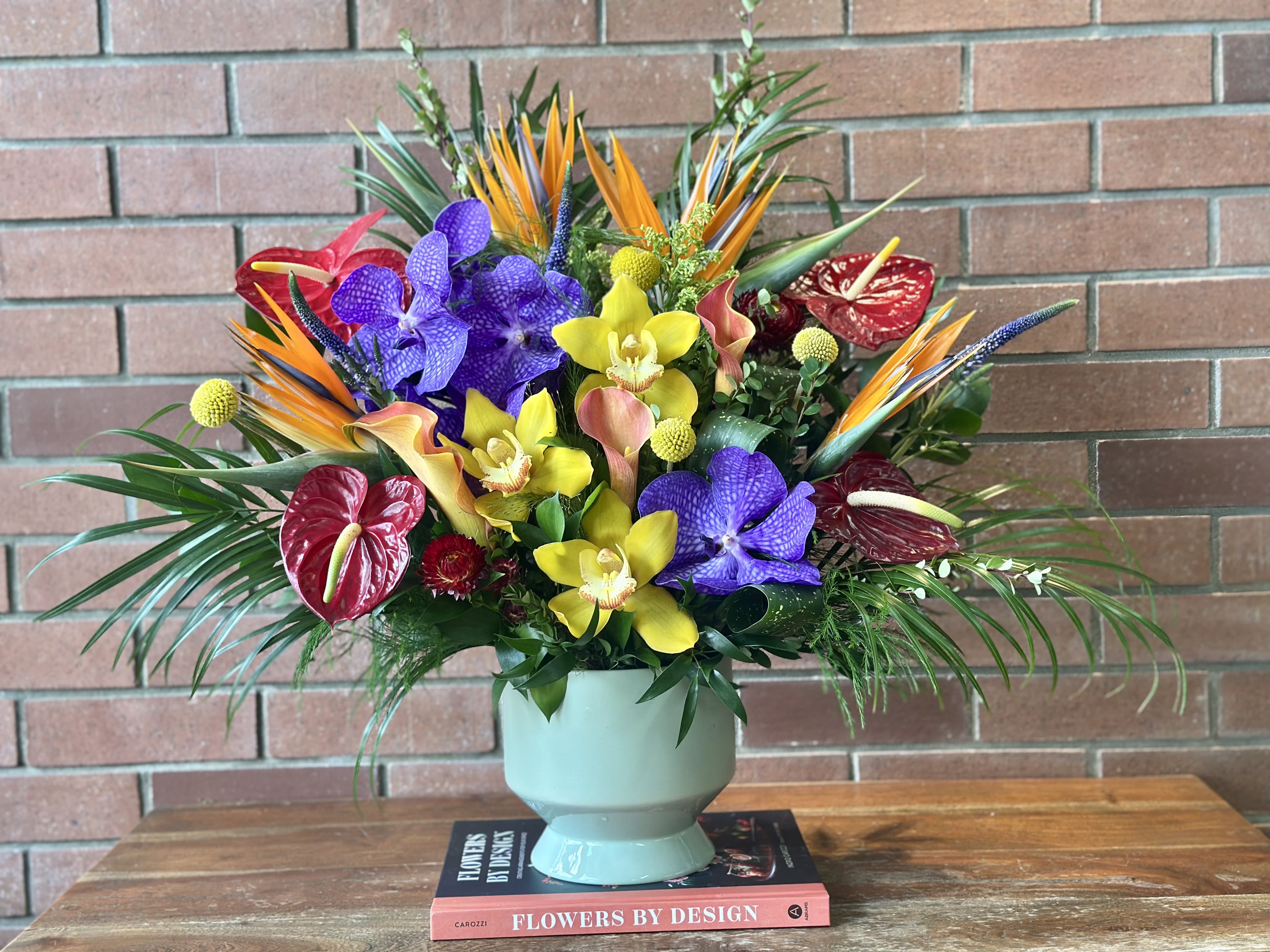 Tall tropical arrangement of orange, purple, yellow, and red flowers in a pale green vase