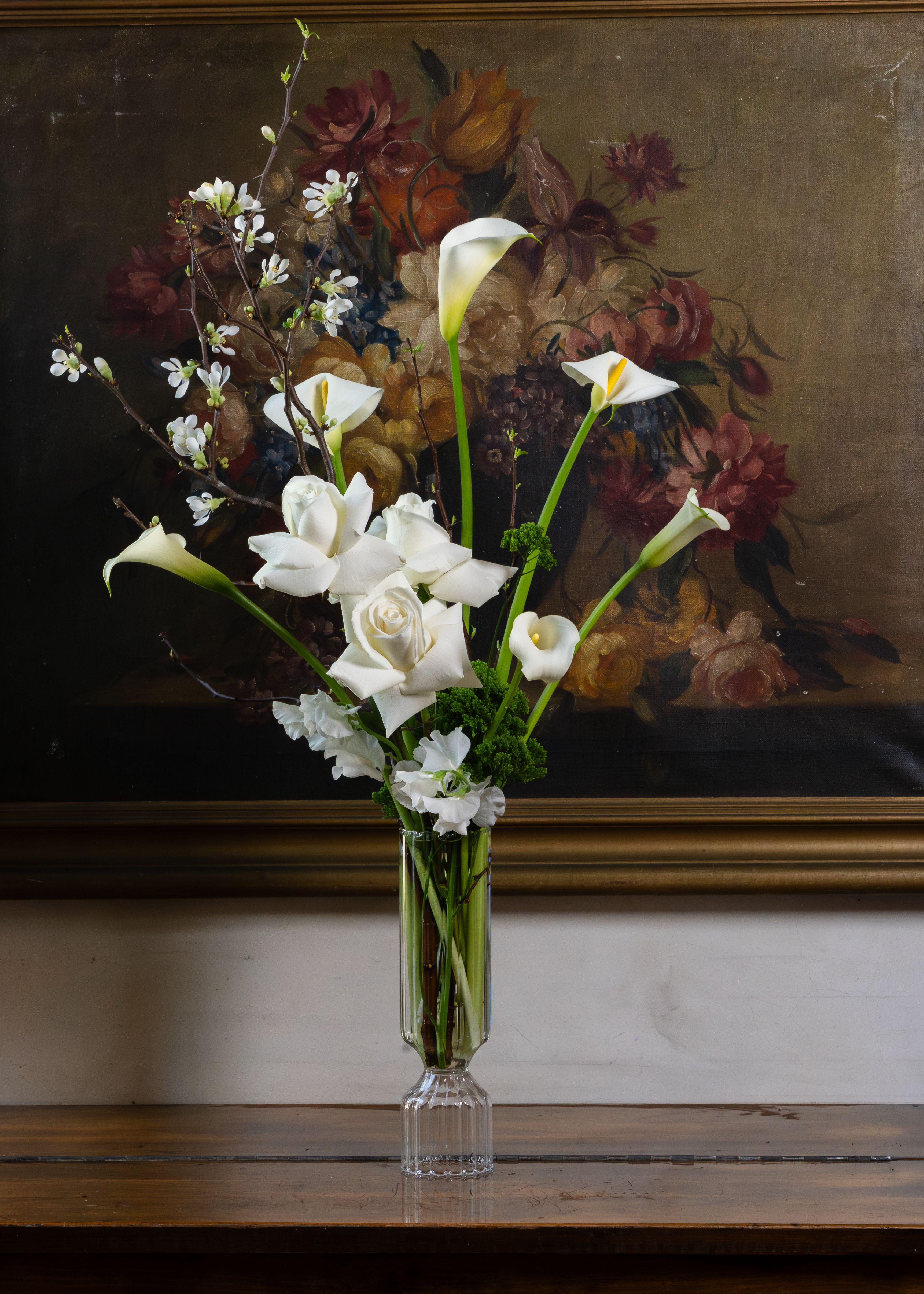 First Blossom - There is something quietly breathtaking about the moment a branch first blooms. That fleeting, delicate instant between winter and spring — that is exactly what this arrangement captures. Presented in a sleek, modern ribbed glass vase, First Blossom is an architectural yet deeply organic composition. Dramatic blooming quince branches reach upward with effortless grace, their tiny white flowers just opening against bare dark wood. Below, sculptural white calla lilies rise on long green stems alongside creamy Playa Blanca roses, flexed open to their fullest, most generous form. White sweet pea adds a soft whisper of delicacy, while lush green trick dianthus grounds the design with rich texture. All white. All premium. Utterly unforgettable.