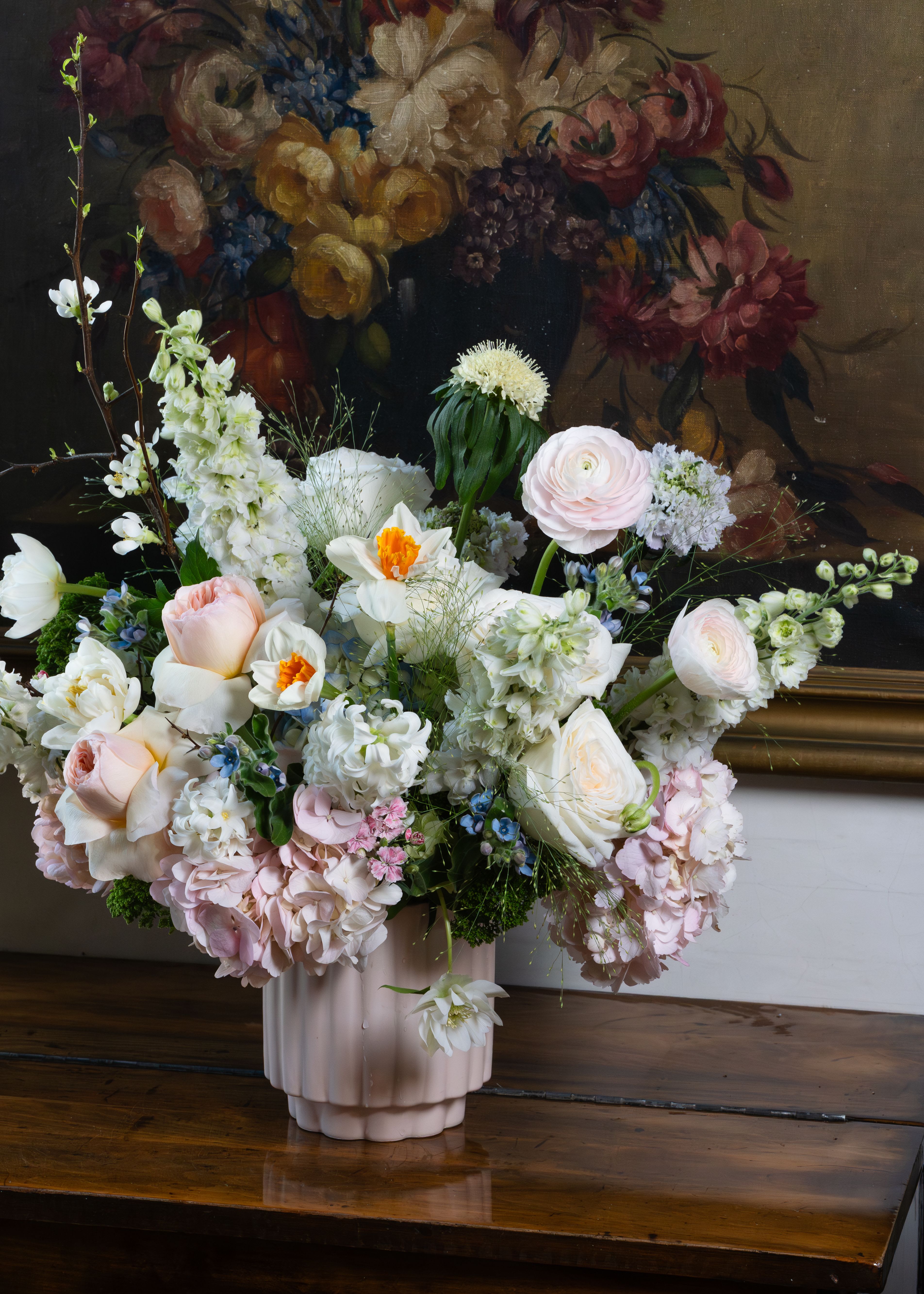 Old Master’s Spring  - Old Master’s Spring Bouquet Some arrangements don’t just hold flowers — they hold a moment in time. Inspired by the lavish floral paintings of the Dutch Golden Age, Old Master’s Spring Bouquet is a breathtaking, abundant composition that feels as though it stepped straight from the canvas. Presented in a soft blush ribbed ceramic vase, this lush garden-gathered design overflows with the finest blooms of the season. Pillowy blush hydrangea cascades generously around the base, setting a romantic foundation for everything that follows. David Austin garden roses in the softest peach and cream open fully at the center, their petals layered like silk. Cheerful daffodils with their signature golden hearts bring a burst of spring sunshine, while delicate pink ranunculus add a tissue-paper softness throughout. White delphinium rises tall, filling the arrangement with its sweet, clove-like perfume. Blooming quince branches reach upward with wild, natural grace, carrying tiny white blossoms on bare woody stems — a reminder that beauty begins long before the garden is in full swing. Touches of blue forget-me-not, pink wax flower, feathery fennel grass, and scabiosa add depth, whimsy, and that effortlessly gathered quality that makes this design feel truly alive. This is spring at its most generous — layered, fragrant, and utterly unforgettable.