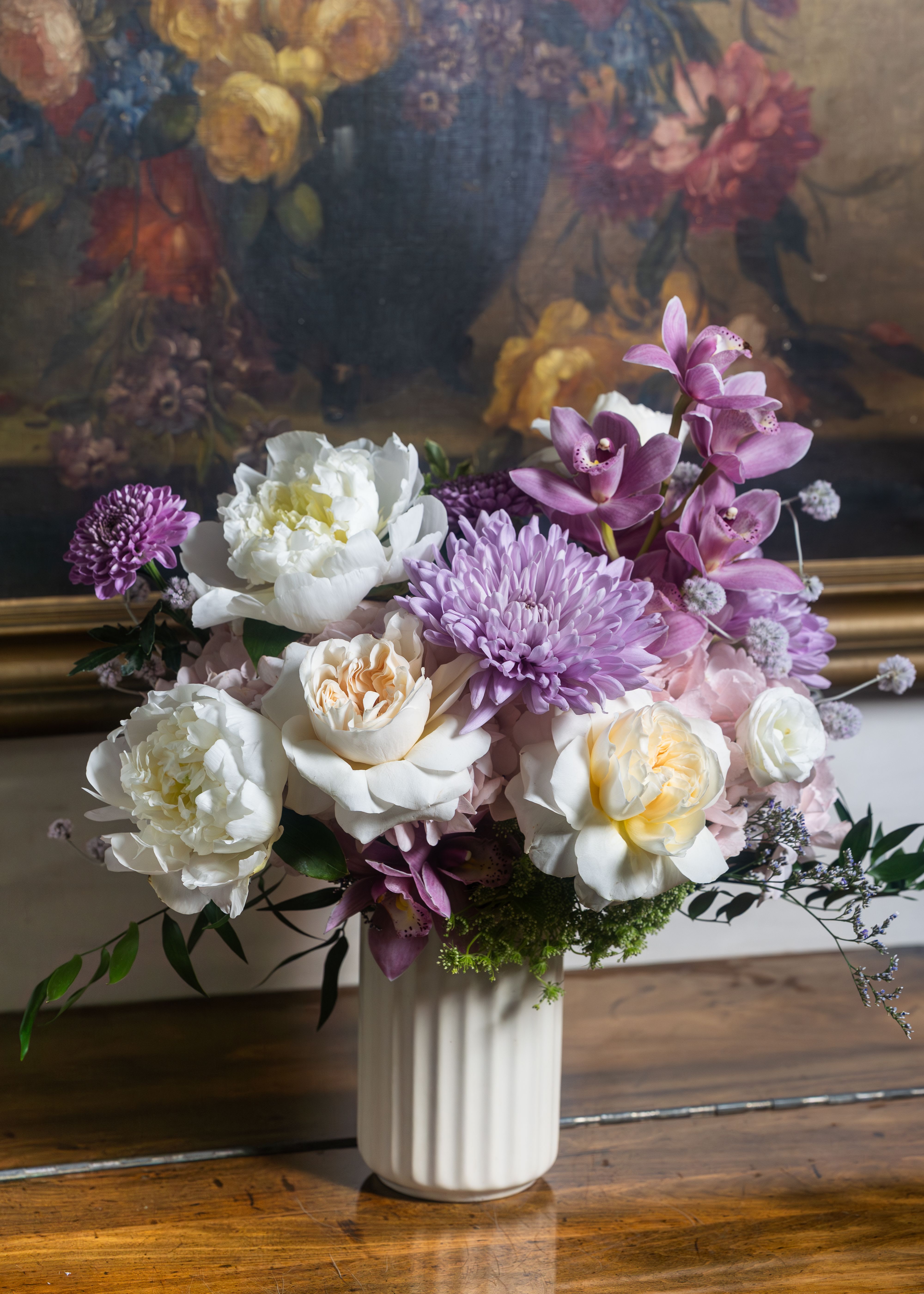 Her Quiet Grace - White peonies open in full, breathtaking bloom — pure, generous, and timeless, just like her. Peach David Austin roses, the world’s most coveted bloom, unfurl in layered perfection at the center. Lavender spider mums spread like a dream across the arrangement, soft and abundant. Purple cymbidium orchids rise with quiet elegance, rare and extraordinary. Blush hydrangea cushions it all in garden softness, while delicate scabiosa and trailing greenery give it the untamed, painterly quality she never even tried to have — it just came naturally. This is not just a bouquet. This is every moment she made beautiful without ever asking for credit.