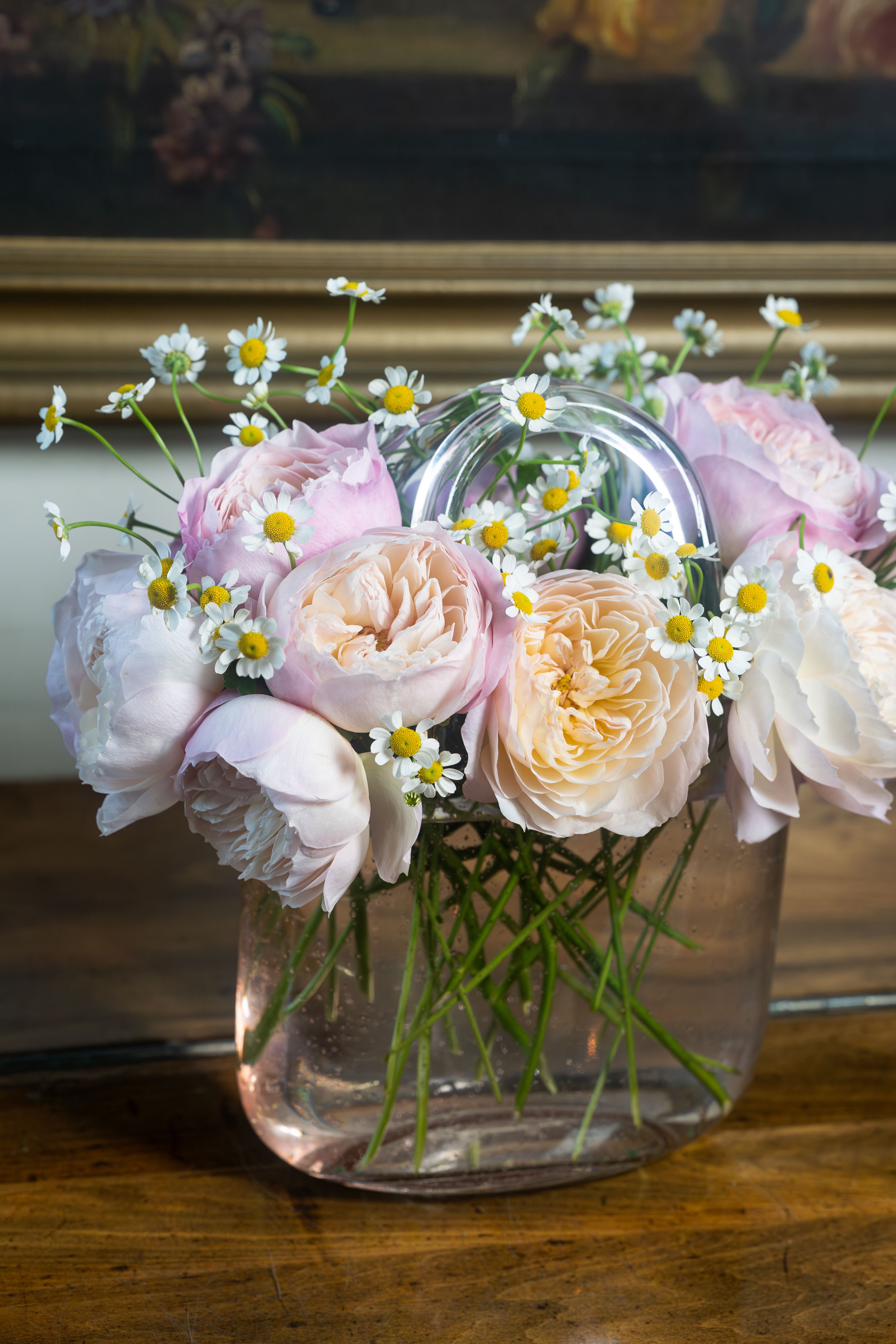 Garden Rose Purse - Soft blush and cream garden roses mingle with pale peach and hints of yellow in a romantic purse-style arrangement-perfect as a birthday bouquet, anniversary gift, or springtime surprise. The soft pastel palette and airy white chamomile with yellow centers give the person you're thinking of a gentle, garden-fresh impression that reads both everyday thoughtful and quietly celebratory.  Large, layered rose blooms sit clustered across the top of a clear square glass vase while delicate daisy-like camomile scatter above and between the roses for lightness and movement. Long stems show through the transparent vase for a freshly cut, casual look; overall size and proportions are ideal for tabletops, mantels, or as a reception centerpiece.  Choose this arrangement for someone who loves soft, feminine floral styling-she'll feel seen and cherished by the delicate color story and scent of garden roses. Vase included; treat with fresh water and a cool spot away from direct sun to keep blooms looking their best.