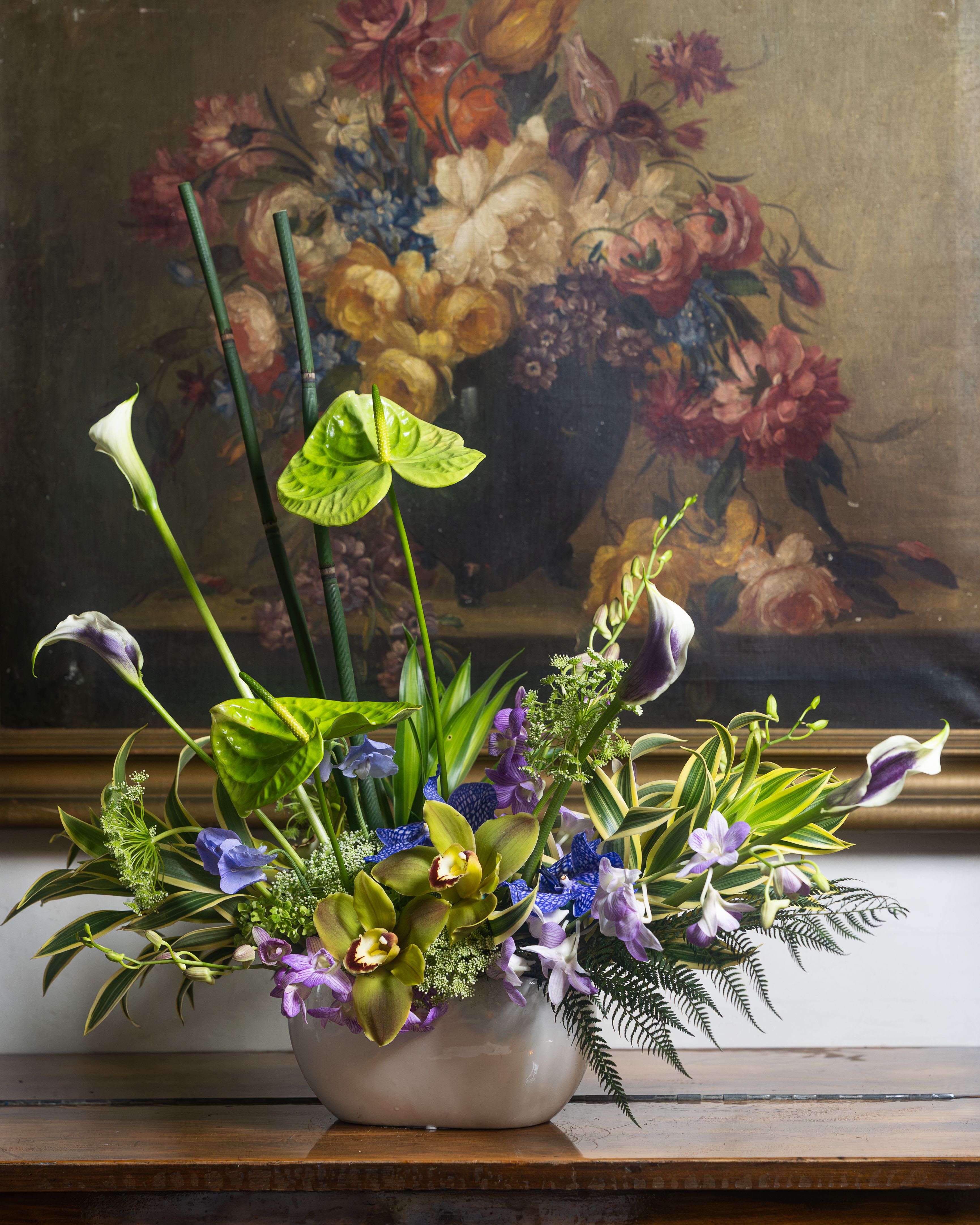 Tropical Opulence - Transport your space to a lush, exotic paradise with this dramatic statement arrangement. Towering bamboo stakes and vivid green anthuriums rise above a richly layered base of chartreuse cymbidium orchids, purple calla lilies, and delicate blue delphiniums — all nestled among cascading variegated dracaena and feathery ferns.  Bold, vibrant, and undeniably luxurious, Tropical Opulence is designed to command attention. Whether gracing an entryway, event table, or living space, this arrangement brings the drama and beauty of the tropics wherever it lands.