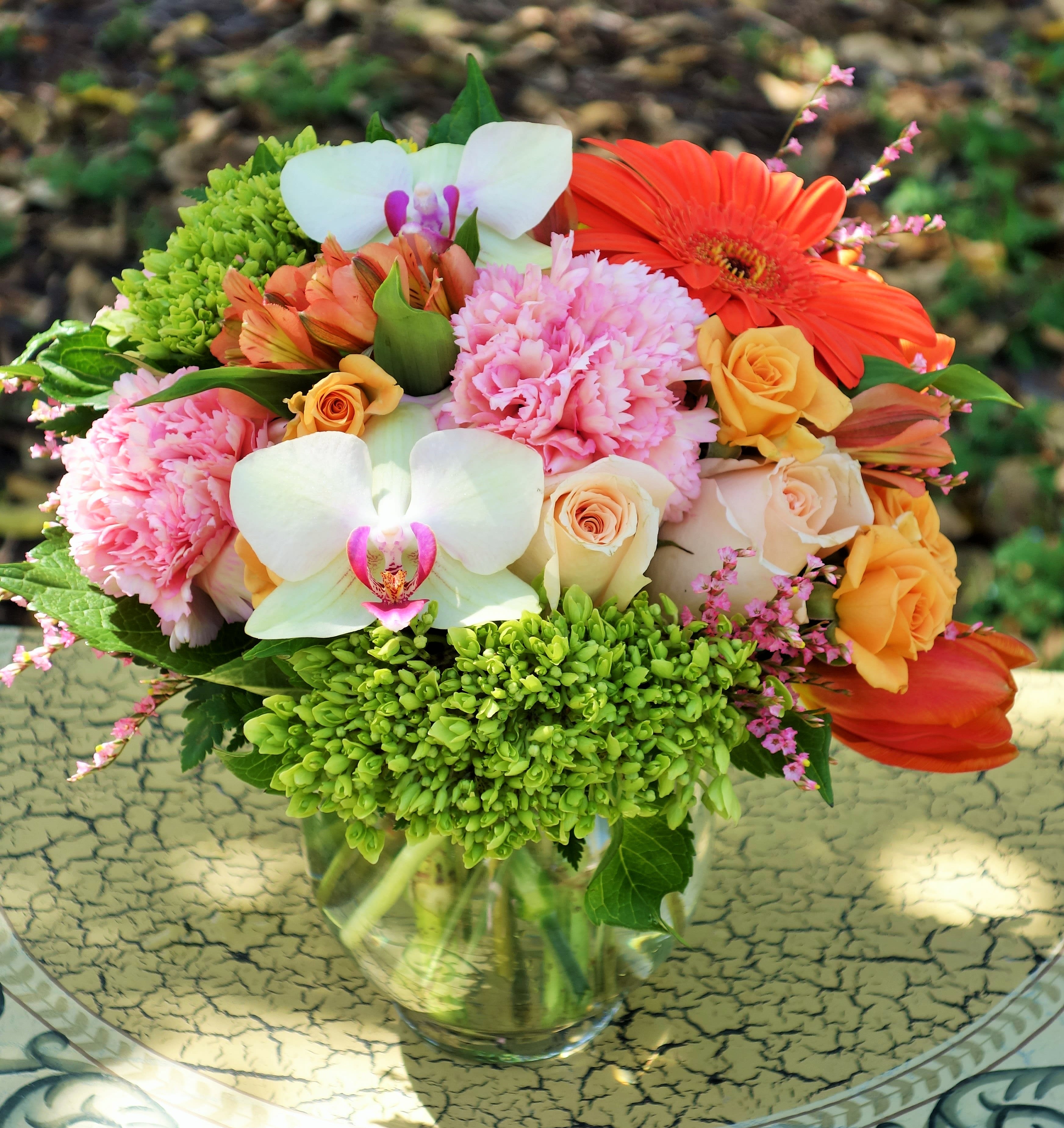 Mixed bouquet of pink carnations, roses, orchids, and orange blooms in a clear glass vase