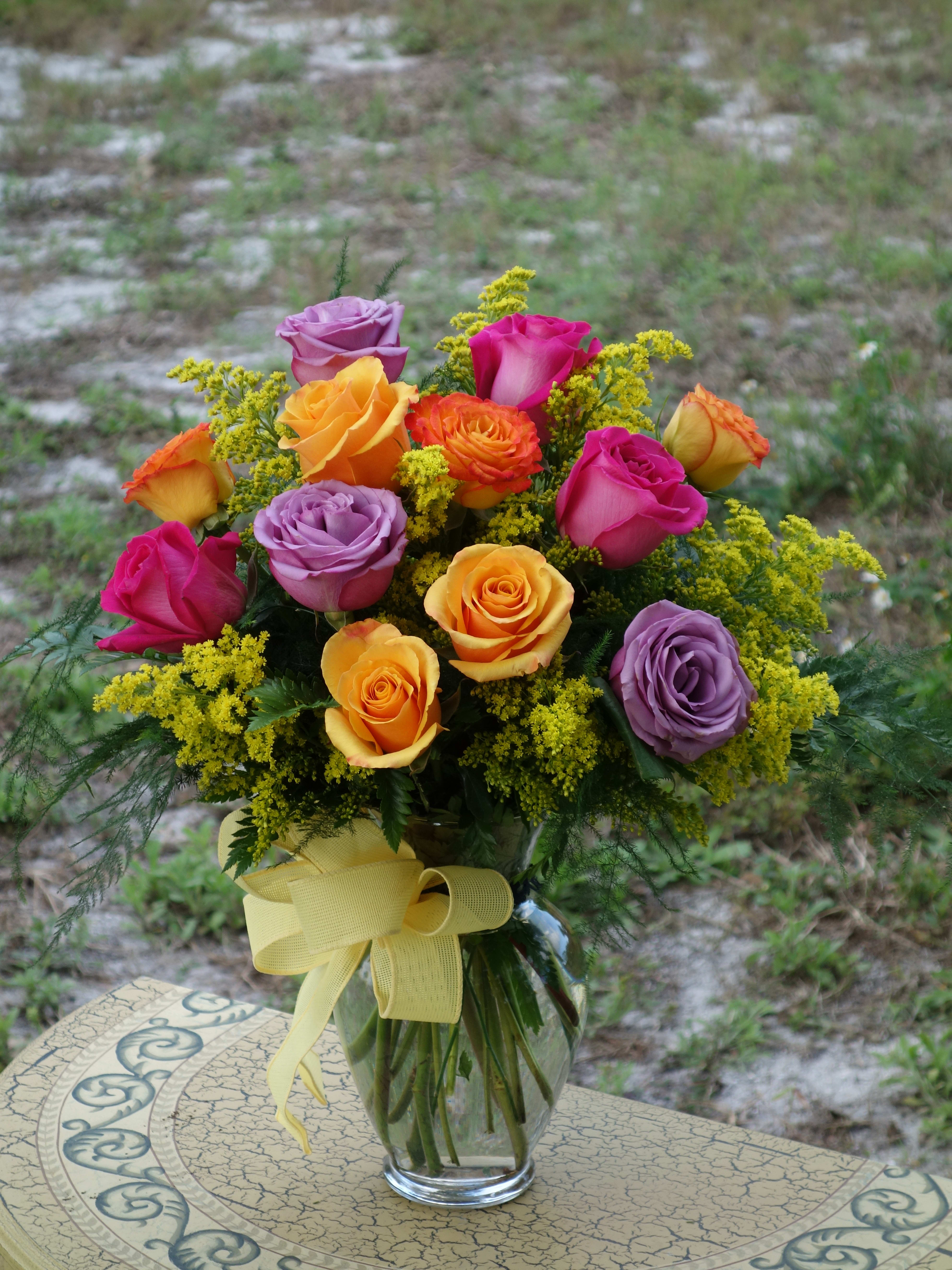 Mixed bouquet of yellow, orange, pink, and lavender roses in a clear vase with a yellow bow