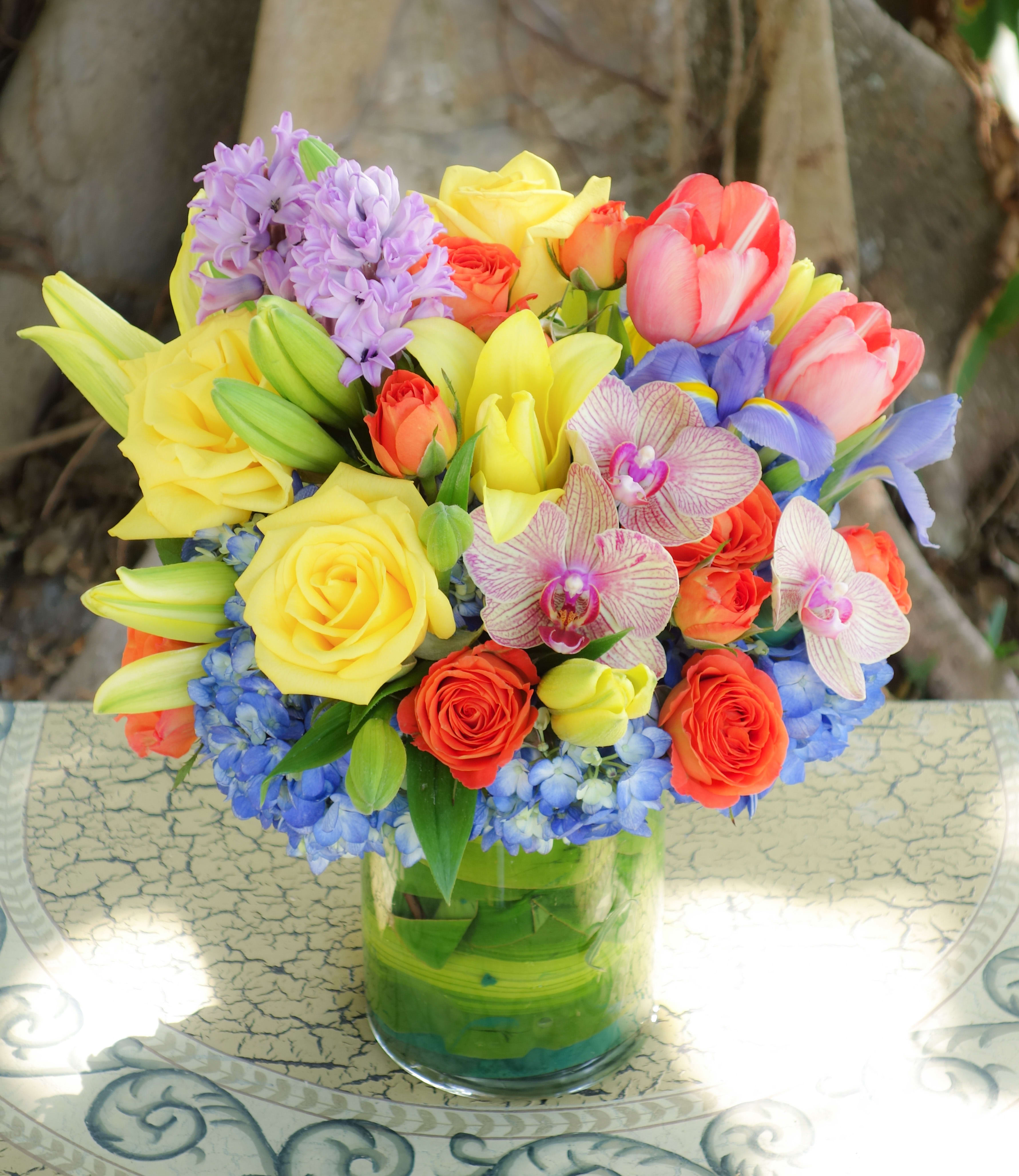 Bright mixed bouquet of yellow roses and lilies with tulips, orchids, and blue hydrangea in a clear glass vase