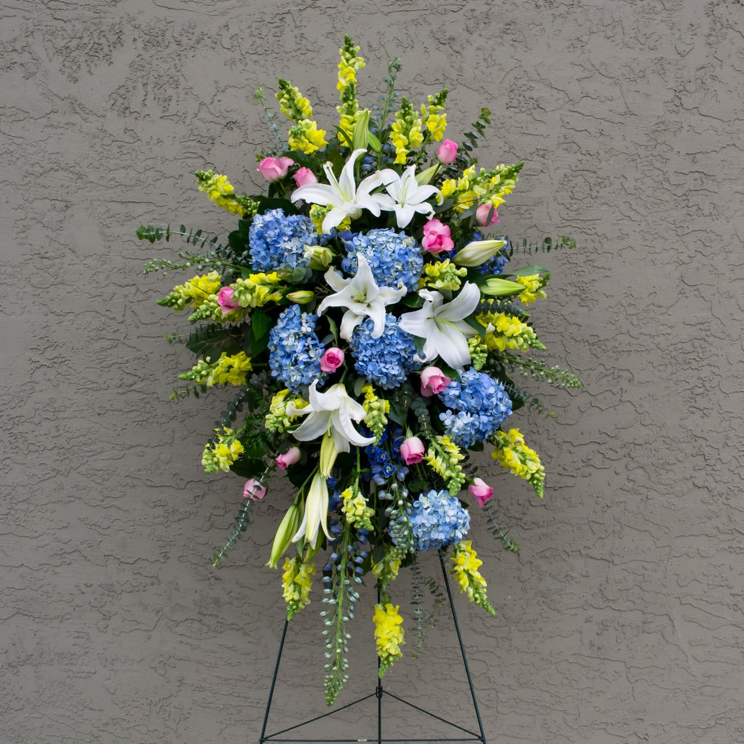 Standing floral spray with white lilies, blue hydrangeas, and yellow snapdragons