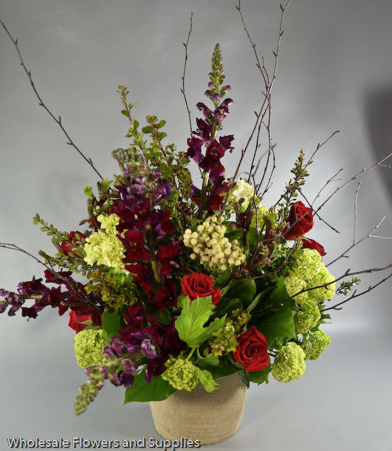 Tall floral arrangement with red roses, purple blooms, and green hydrangeas in a burlap-wrapped vase