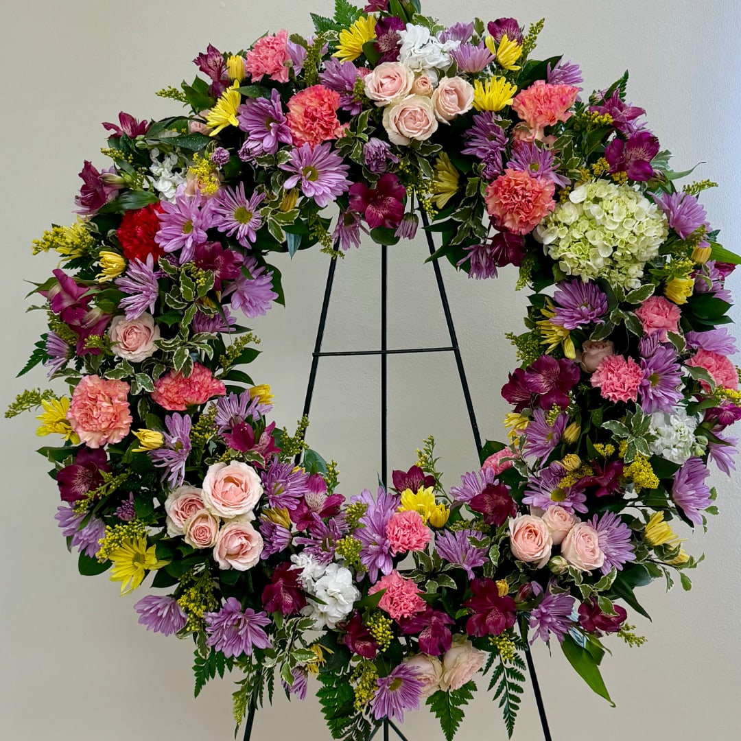 Heart-shaped floral wreath with pink, purple, yellow, and white blooms on a black stand