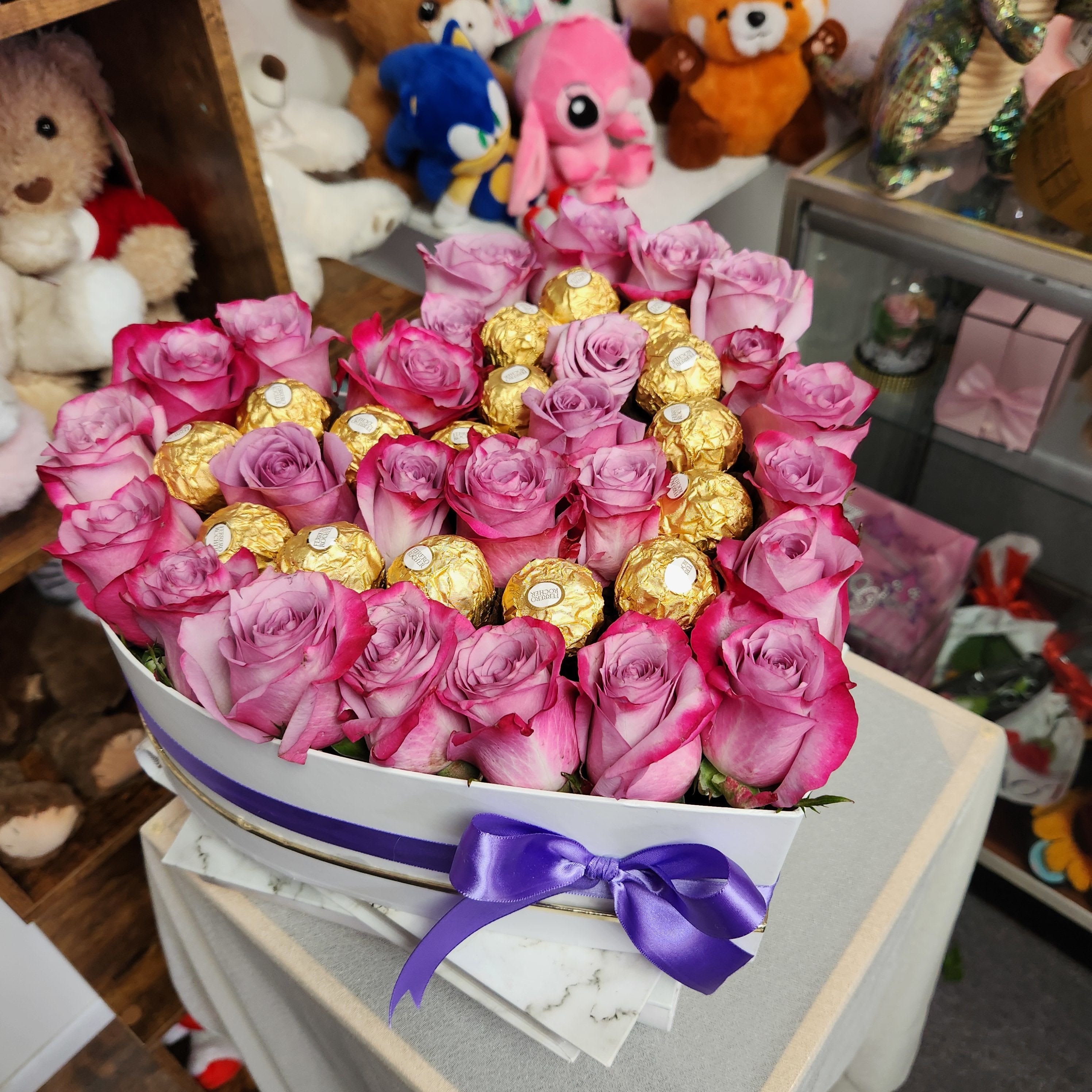 Pink roses arranged with gold-wrapped chocolates in a white box with a purple ribbon