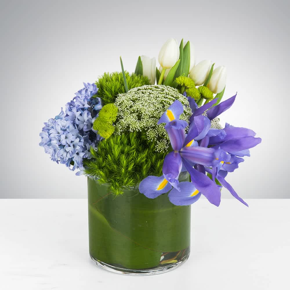Mixed bouquet of white tulips, purple irises, and blue flowers in a green glass vase