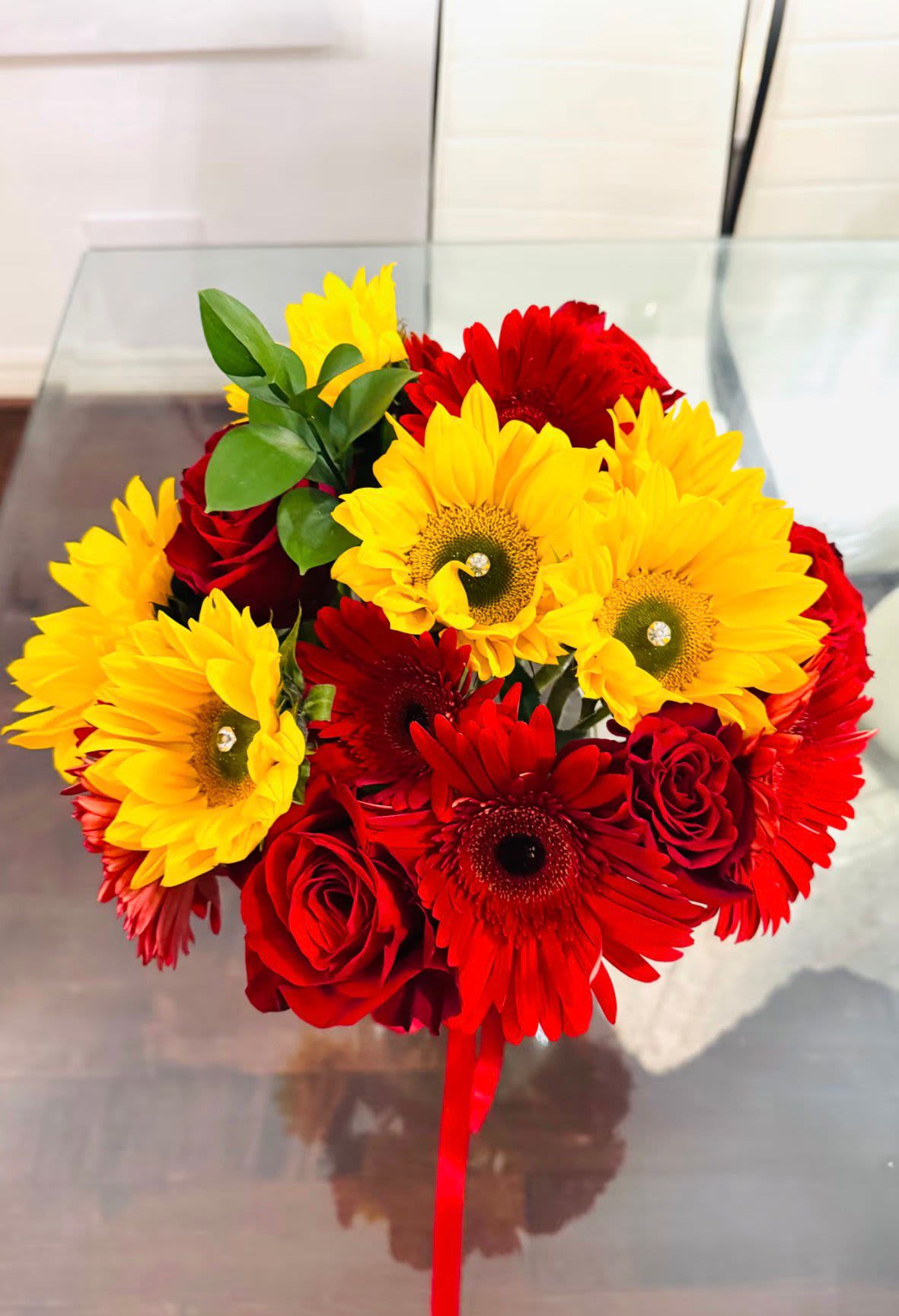 Joy and Grace Gerbera with roses - This Bouquet is a striking composition of romantic fire and golden light, blending the timeless allure of red and pink roses with the radiant joy of sunflowers, and Gerbera. Each bloom plays its part in a beautiful contrast- the roses deep crimson, speak of devotion, desire, and enduring love. 