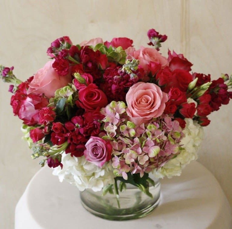 Round arrangement of pink and red roses with hydrangeas and stock in a clear glass cylinder vase