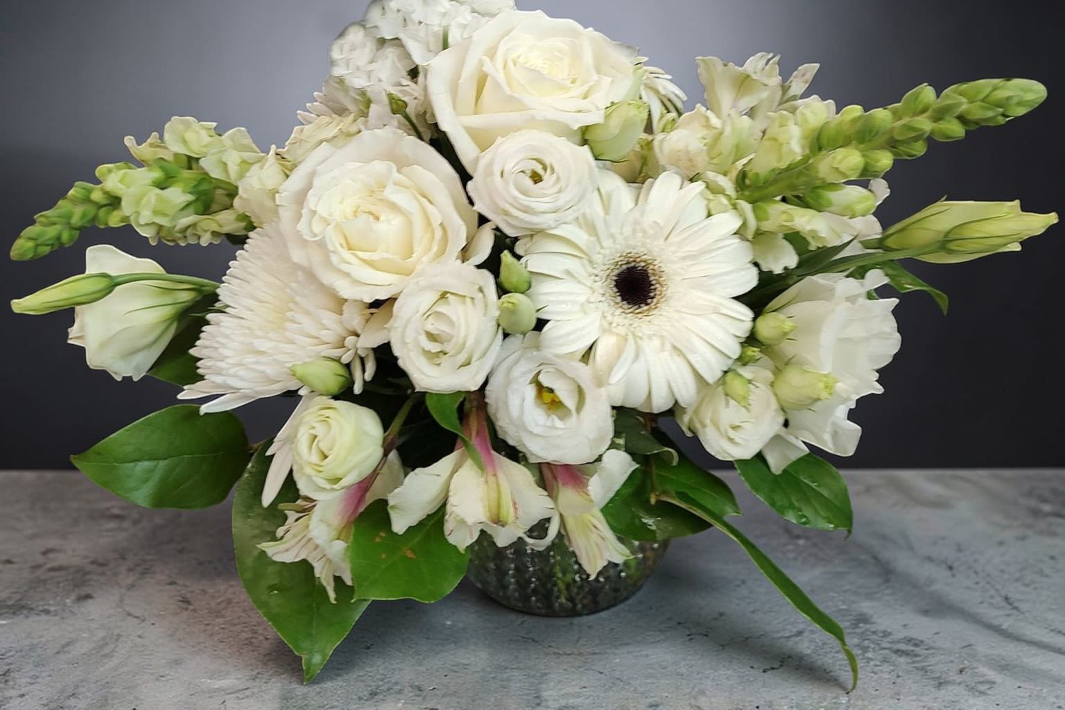 Ivory Whisper - This is a soft, elegant floral arrangement built around a serene all-white palette with touches of fresh green. It has a rounded, slightly asymmetrical shape that feels natural yet refined.  The design features a mix of blooms with varied textures:  Full, layered white roses and ranunculus create a lush, romantic core A striking white gerbera daisy adds a bold focal point with its dark center Delicate lisianthus and possibly snapdragons introduce height and gentle movement Subtle greenery with broad, glossy leaves frames the arrangement and gives it balance  The flowers are arranged in a low, rounded glass vase, making it suitable as a centerpiece. The overall effect is clean, calming, and timeless—perfect for weddings, formal events, or minimalist interiors.