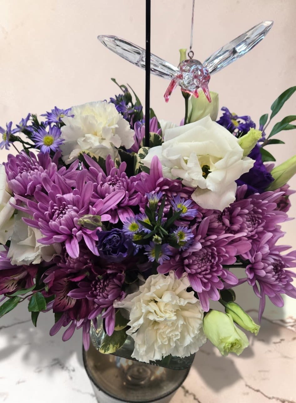 Art and Flowers Exclusive "Whispers Of Spring" - A compact cluster of soft purples, pinks, and creams-this flower arrangement "Whispers Of Spring" offers a gentle, uplifting presence perfect for a birthday, anniversary, housewarming, or a thoughtful thinking-of-you gesture. The low, rounded design and delicate hummingbird ornament create a keepsake focal point that feels both celebratory and quietly comforting, also appropriate for sympathy or a serene Easter display.  Each arrangement is thoughtfully gathered in a clear glass cube vase (or clear glass vase depending on the version), with purple chrysanthemums and lavender asters forming the colorful mound, creamy white carnations and pale green lisianthus adding ruffled texture, and pink roses lending a soft, romantic touch. A faceted crystal or blue glass hummingbird-mounted on a slender stake or pick-rises above the blooms to catch the light and add a whimsical, lasting accent. The compact size makes it perfect for tabletops, desks, or mantel displays and suits seasonal spring gifting as well as year-round occasions like Women's Day, Best Friends Day, or Admin Professionals' Week.  Designed to make the recipient feel seen and comforted, this arrangement sends a message of warmth and gentle celebration whether you're marking an anniversary, welcoming someone into a new home, or offering sympathy. Care is simple: keep water fresh and display out of direct hot sun to preserve the blooms; the glass hummingbird makes it a small keepsake to enjoy after the flowers fade.