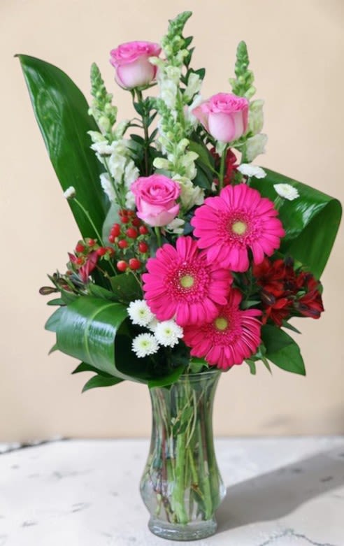 Blushing Garden Elegance by Art and Flowers -  This vibrant floral design showcases a lively mix of pinks, whites, and lush greens arranged in a tall, clear glass vase. Soft pink roses rise gracefully above the arrangement, adding height and refinement, while bold pink gerbera daisies create a cheerful focal point at the center. White snapdragons and petite white filler flowers provide vertical movement and light contrast. Glossy, sculpted green leaves frame the bouquet with a modern, architectural touch, and accents of  berries add depth and texture. Overall, the design feels fresh, uplifting, and contemporary—perfect for celebrations, congratulations, or simply brightening a space. Colors may vary due to availability. 