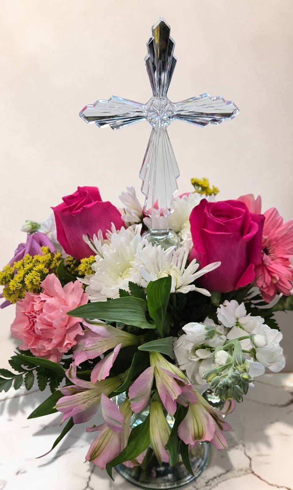 Heavenly Grace - A graceful arrangement featuring a crystal cross surrounded by pink roses, white chrysanthemums, carnations, and alstroemeria. Soft pastel tones and lush greenery create a peaceful, uplifting design full of elegance and heartfelt beauty.