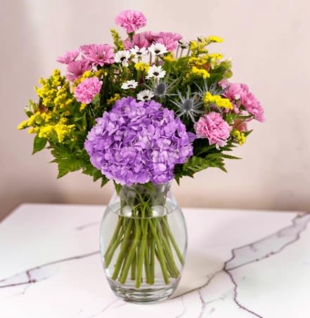 Spring Harmony  -  A colorful bouquet of flowers arranged in a textured purple glass vase. The bouquet features a mix of blooms, including soft purple hydrangeas at the center, pink carnations and daisies, small white flowers, and clusters of bright yellow filler flowers, along with green foliage. This design will create a mood that feels cheerful, fresh, and decorative, like a floral still life.
