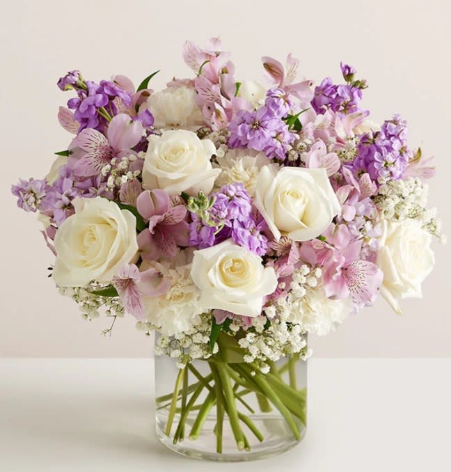 Lavender Whisper by Art and Flowers - This arrangement is a soft, romantic blend of pastel elegance and airy texture. Creamy white roses serve as the focal point, their full, classic blooms offering a sense of timeless beauty. They are surrounded by delicate lavender stock and blush-pink alstroemeria, adding gentle movement and a light, garden-fresh feel. Clusters of white filler flowers create a cloud-like base, enhancing the softness and giving the design a lush, rounded shape. Arranged in a clear glass cylinder, the visible stems add a clean, modern touch that balances the arrangement’s romantic charm. 