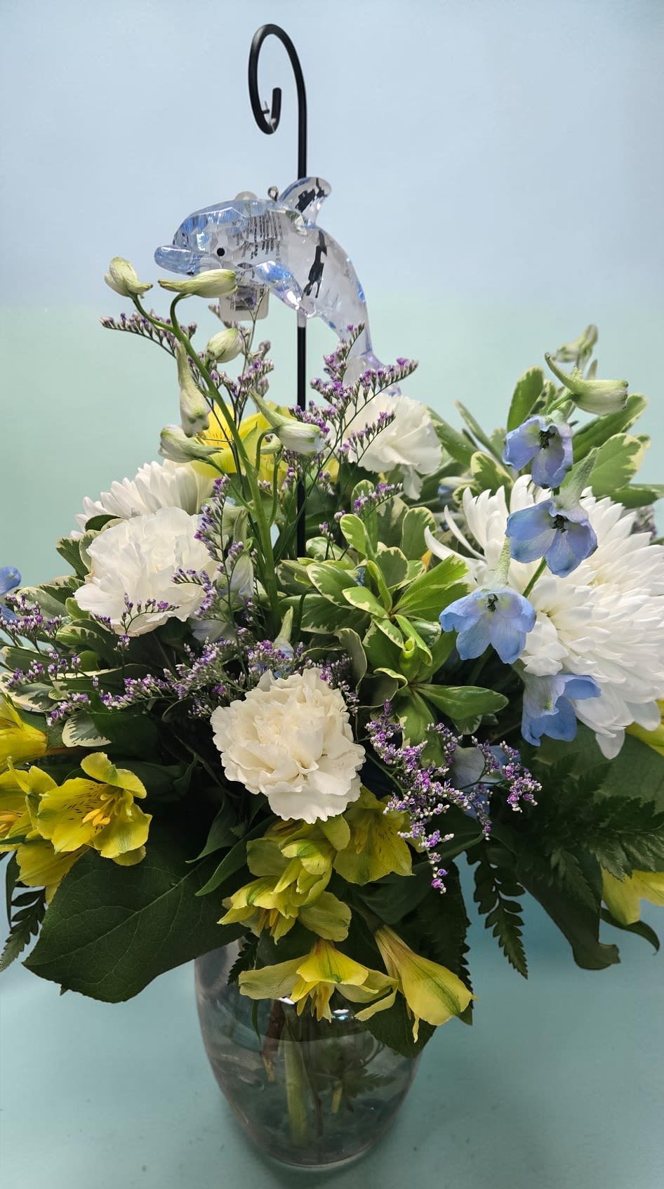 Ocean Whisper---Four versions Available - A whimsical fish ornament with iridescent blue and silver scales, suspended above a vibrant floral arrangement in a glass vase. The arrangement features white chrysanthemums, yellow alstromeria, and delicate blue flowers, with lush green foliage.   