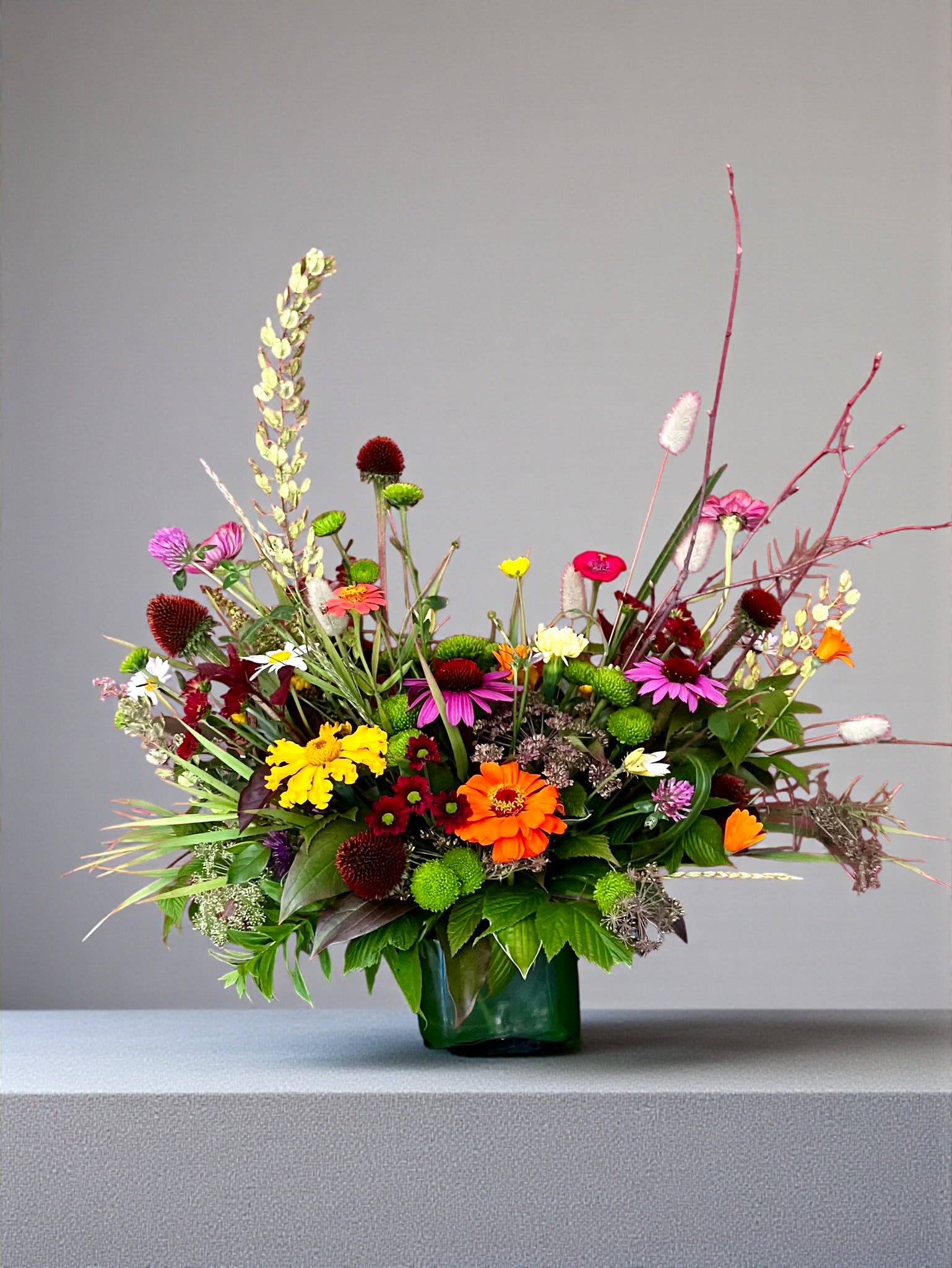 Rustic Spring Serenade -  Rustic Spring Serenade--Flemish Design Style  This whimsical garden-style arrangement is a joyful celebration of spring’s playful beauty. Designed in a simple natural wooden box, it features many of Spring's best blooms: cheerful golden gerbera daisies dancing among delicate pink ranunculus, lavender tulips, blush spray roses, and soft white accent blooms. Airy textural greenery and trailing botanical elements create movement and an organic, free-flowing silhouette. Each design will vary due to availability and seasonal products.   The asymmetrical design, Flemish Style, gives it a fresh-from-the-garden feel, while the vibrant contrast of sunny yellows and romantic pinks brings warmth and charm. With its loose, artistic composition and rustic container, this arrangement feels both modern and naturally elegant—perfect for brightening a space with effortless springtime sophistication.