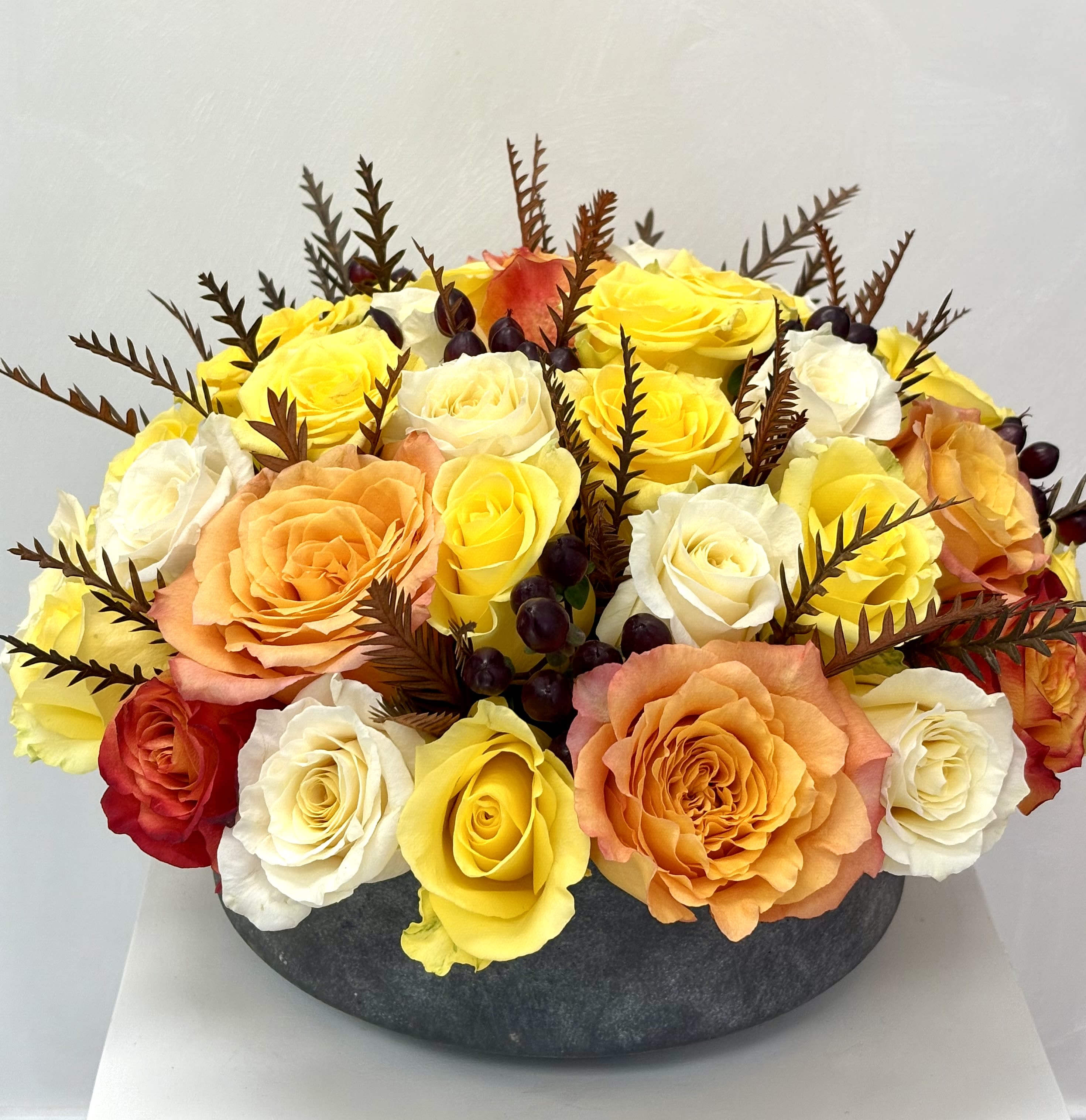 Round arrangement of yellow, white, orange, and red roses in a dark vase