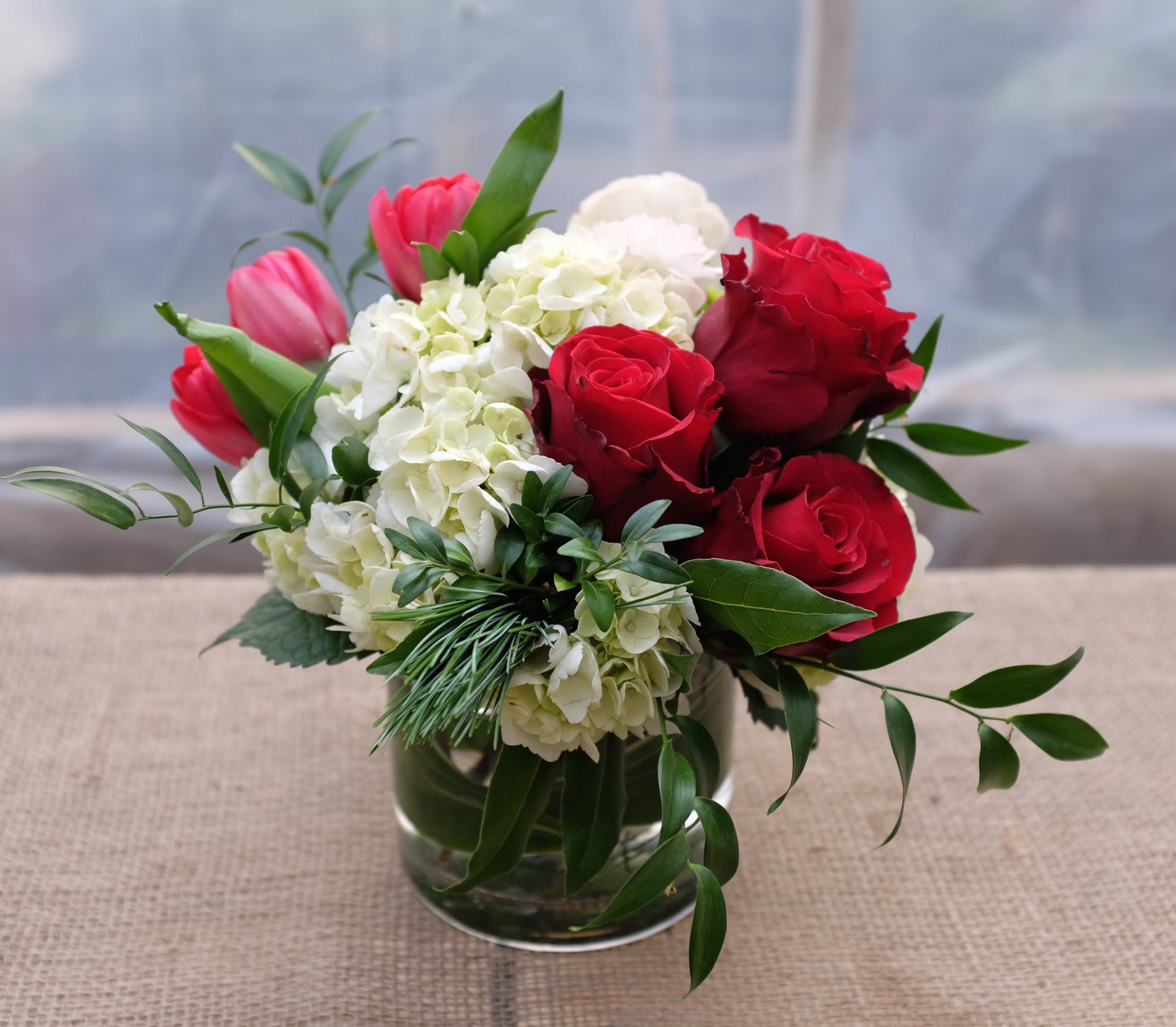 Red roses and pink tulips arranged with white hydrangeas in a glass vase.