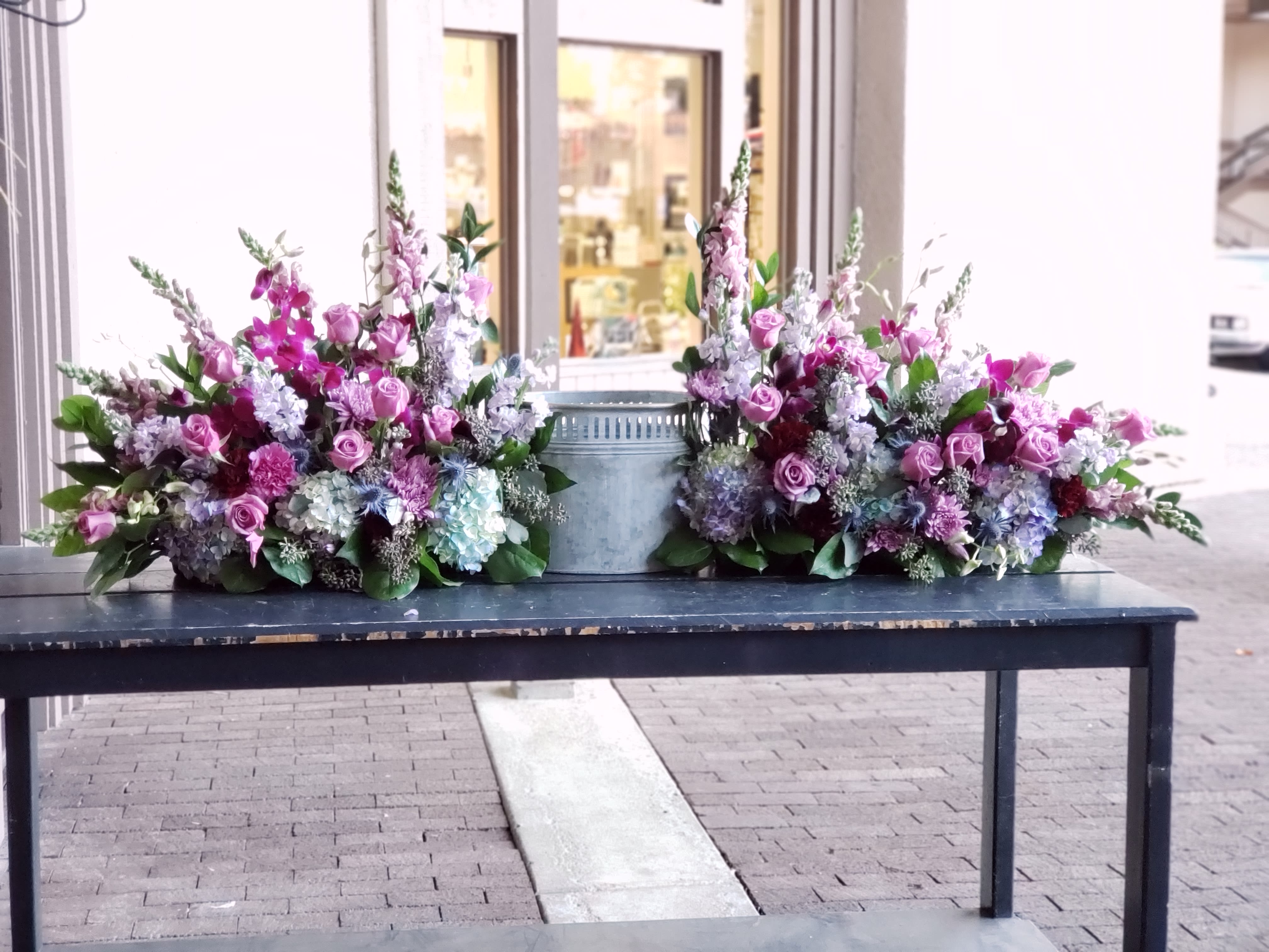 Two pink and lavender floral sprays flank a metal urn on a table