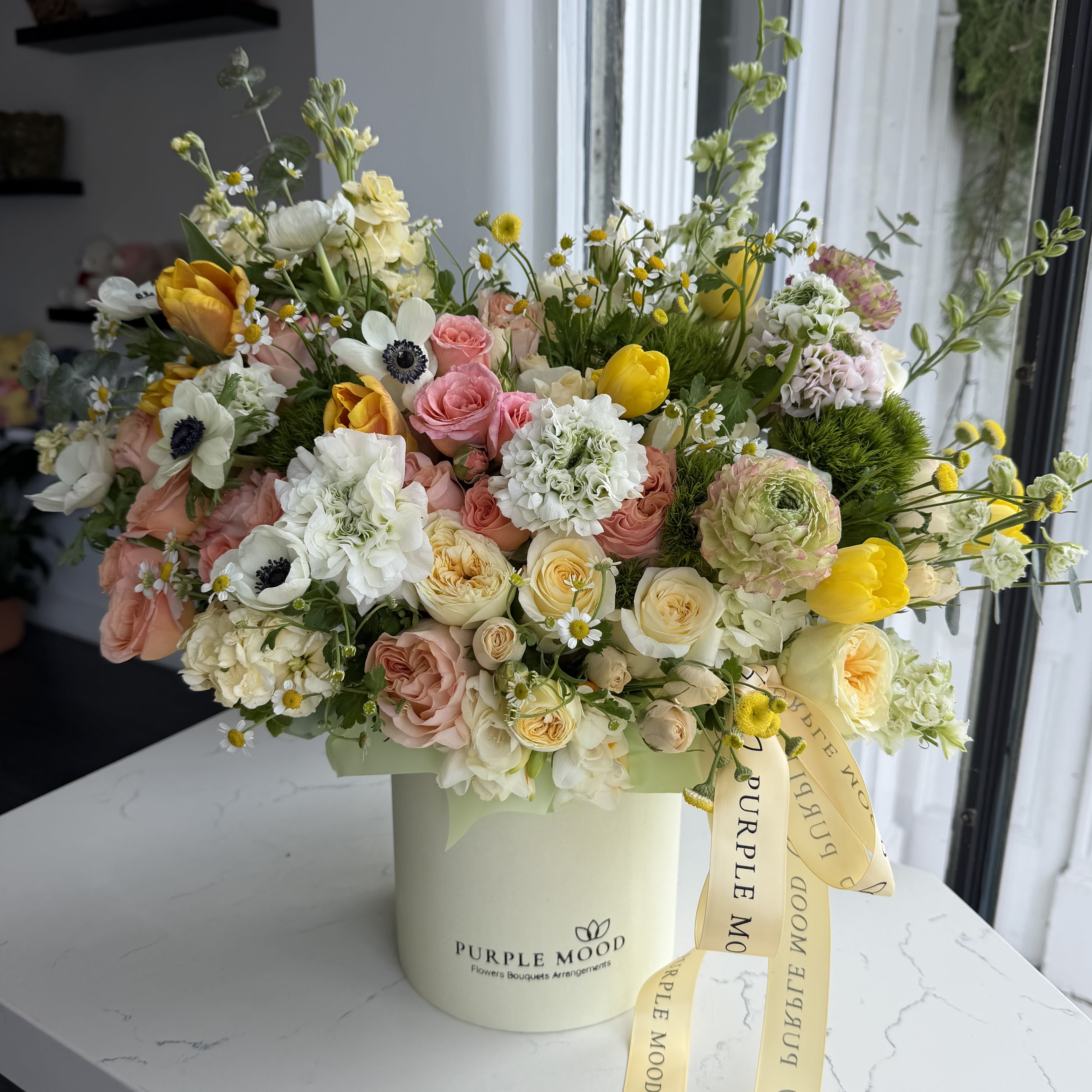 Sunny Meadow Box - A vibrant floral meadow in a box! Sunny Meadow Box features fresh greens, soft peach roses, delicate anemones, and cheerful yellow accents, creating a lush and lively arrangement. Inspired by a sunlit meadow, this design brings warmth, joy, and natural beauty to any space.