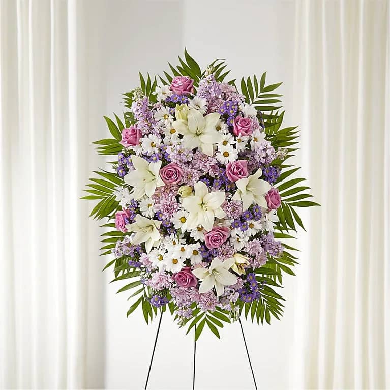Blush Halo - A soft, oval standing spray designed to offer gentle comfort and quiet beauty-Blush Halo is a sympathy standing arrangement that reads as both formal and tender. Pink roses and white lilies form the luminous core so the person you're thinking of will feel remembered; clusters of lavender and purple filler flowers and small white daisy-like blooms add delicate texture while broad palm fronds frame the composition for a respectful, ceremonial presence.  This large, densely packed arrangement is mounted on a black easel and shaped into a rounded, wreath-like mass that suits memorial services, funerals, or a serene celebration of life. The palette of pink, white, purple, and green creates a calm, romantic mood without feeling flashy; blossoms include roses, lilies, daisies, asters, chrysanthemums, and other seasonal filler flowers. Order with confidence for a meaningful tribute-the standing easel ensures visibility at services, and the classic, symmetrical styling makes this arrangement appropriate whenever you need a dignified, comforting floral expression.