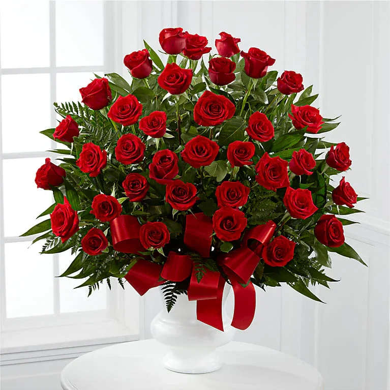 Red Pinnacle - A lush dome of deep red roses makes an immediate impression-the perfect anniversary roses or Valentine's bouquet that reads as classic romance. This flower arrangement places densely packed red roses in a rounded, upright form so the recipient is met with a bold wall of blooms; the design is equally right for marking a milestone, saying "I love you," or bringing everyday luxury to someone special.  Each bloom is nestled against glossy, dark green foliage and presented in a clean white ceramic pedestal vase, the contrast sharpening the roses' rich color. A wide red satin ribbon tied around the base adds a polished, celebratory finish and reinforces the romantic palette of red, green, and white. The arrangement is formal in silhouette yet warm in feeling-designed to sit beautifully on a dining table, mantle, or bedside.  Choose this for anniversaries, Valentine's Day, or any love-themed gifting moment when you want the person you're thinking of to feel treasured and seen. The compact, rounded presentation keeps stems secure in the white vase for easy display; place in fresh water and away from direct heat to enjoy peak bloom for as long as possible.