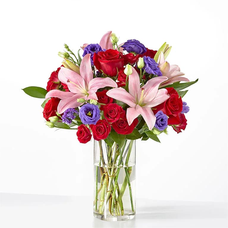 In Full Contrast - Striking red roses meet soft pink lilies in a full, rounded mixed arrangement that reads as lush and romantic - perfect as a birthday bouquet, anniversary flowers, or a heartfelt way to say "I'm thinking of you." Purple lisianthus and other lavender-toned accents deepen the color story, adding contrast and a celebratory, spirited feel the recipient will notice the moment it arrives.  This dense, layered design is gathered in a clear cylindrical glass vase so the stems are visible for a classic florist presentation. The palette of red, pink, purple and white creates visual depth and texture: velvety roses, delicate lisianthus, and elegant lilies combine for a fragrant, abundant display that fills a room.  A versatile everyday arrangement, it's the kind of gift that makes someone feel truly seen - ideal for anniversaries, moments of romance, birthdays, or simply to say thank you. Vase included; arrange delivered ready to display with simple care (fresh water and trim stems every few days) to keep blooms looking their best.