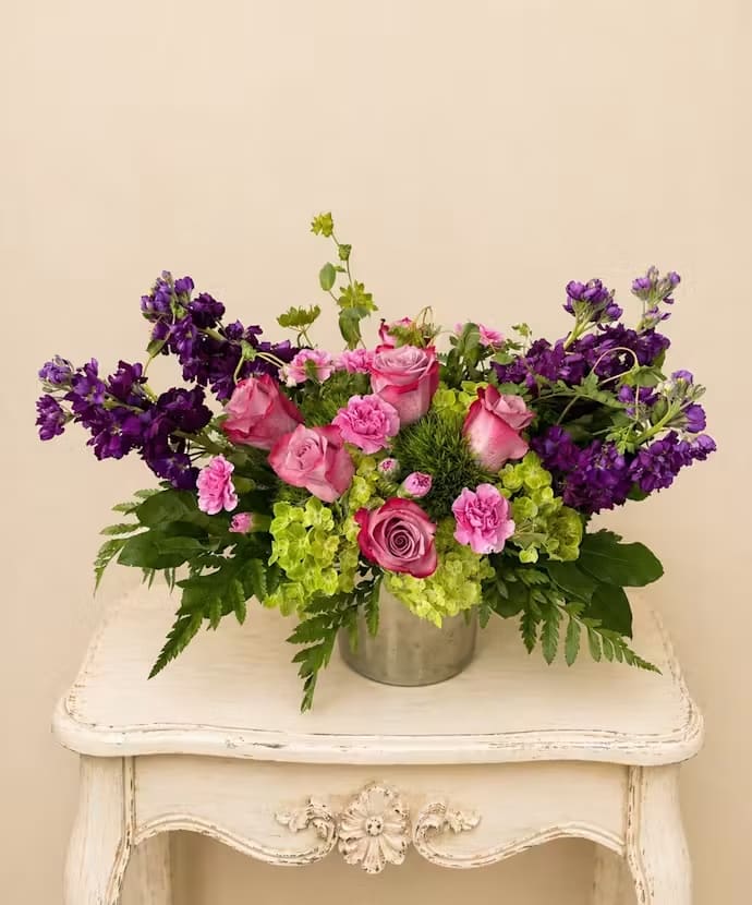 Pink After Dark - Luminous pink roses and carnations mingle with vivid purple blooms and chartreuse-green hydrangea-like clusters in a wide, low bouquet designed for everyday celebration. This pink and purple floral arrangement is perfect as a birthday bouquet, a cheerful anniversary gift, or a just-because surprise that tells someone you're thinking of them in a joyful, romantic way.  Set in a simple metal container that lets the flowers spill outward, the design reads lush and abundant without being fussy-bright pinks, deep purples, and lime-green accents create lively contrast and a garden-fresh texture. The mix includes roses for classic romance, carnations for lasting color, and voluminous green florets to add fullness and modern pop; its low profile makes it an ideal centerpiece for a table or sideboard.  The person you send this to will feel genuinely celebrated by the arrangement's bold color play and generous shape. Arrives ready to display in the included metal container; keep in fresh water, trim stems occasionally, and place away from direct heat to extend vase life.
