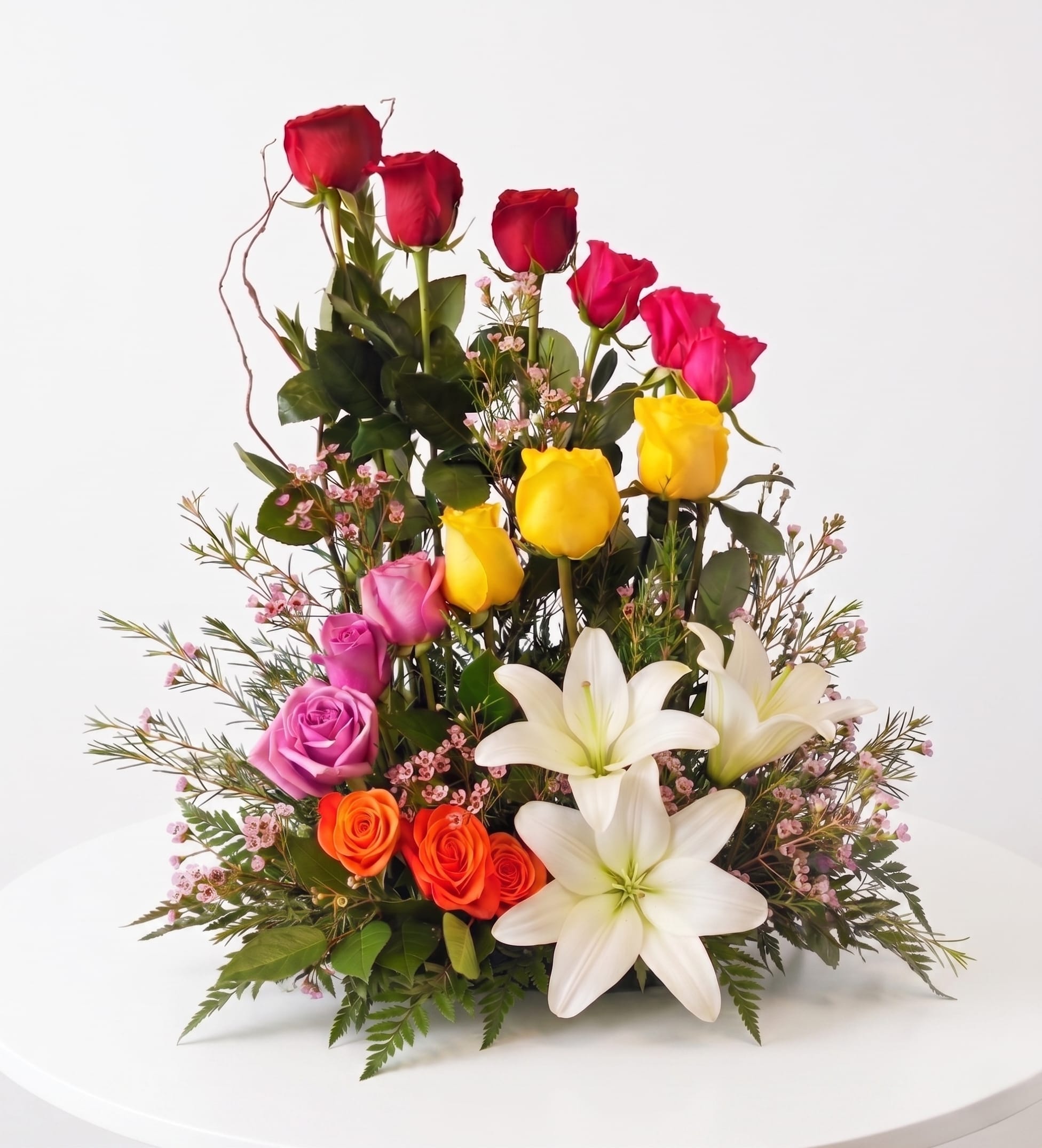 River Walk Fiesta - Bring the vibrant energy, color, and celebration of San Antonio straight to their door! Inspired by the festive spirit of our famous River Walk, this showstopping arrangement features a dramatic, rising cascade of premium roses in a full spectrum of colors—from fiery orange and deep crimson to sunny yellow, hot pink, and lavender.   Anchored by large, pristine white lilies and accented with delicate waxflower, this bold, tiered design is the ultimate way to celebrate birthdays, milestones, or any moment that calls for a little extra joy.   Perfect For: Birthdays, Get Well Wishes, Anniversaries, Congratulations, "Just Because," or a stunning local centerpiece.
