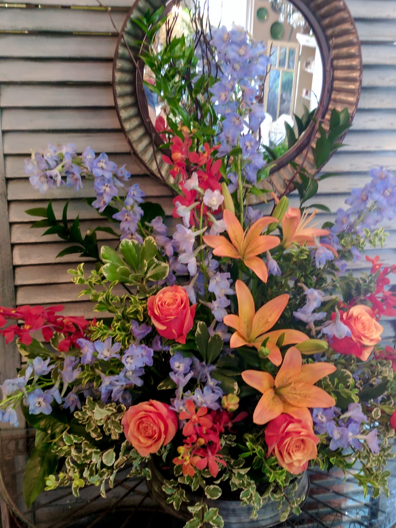 Tall arrangement of peach lilies, coral roses, blue delphinium and red orchids in a metal container