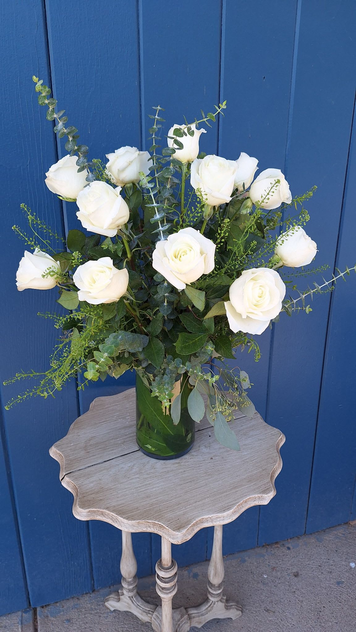 Tall arrangement of white roses with filler in a clear glass vase on a small wooden table