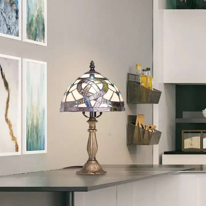 Celtic Pathways Tiffany Style Memory Lamp - A domed Tiffany-style memory lamp that brings handcrafted vintage charm to any room - perfect as a thoughtful housewarming gift, sympathy keepsake, or special birthday present for someone who loves classic decor. The table lamp reads like a work of art: the stained-glass shade blends mostly cream panes with rich purple and blue accents and dark mosaic details that glow when lit, creating a soft, comforting atmosphere for bedside tables or living room sideboards.  The lamp sits on an ornate warm-toned metal pedestal that adds traditional, slightly antique character and a sturdy presence. The shade's mosaic pattern and domed silhouette cast textured pools of color and light, making it as much a decor focal point as a functional light source. Sized for tabletops, it's a versatile piece for interior styling and keepsake gifting.  Give this to the person you want to soothe, remember, or celebrate - it's the kind of gift that says "I'm thinking of you" and keeps giving warmth every evening. 