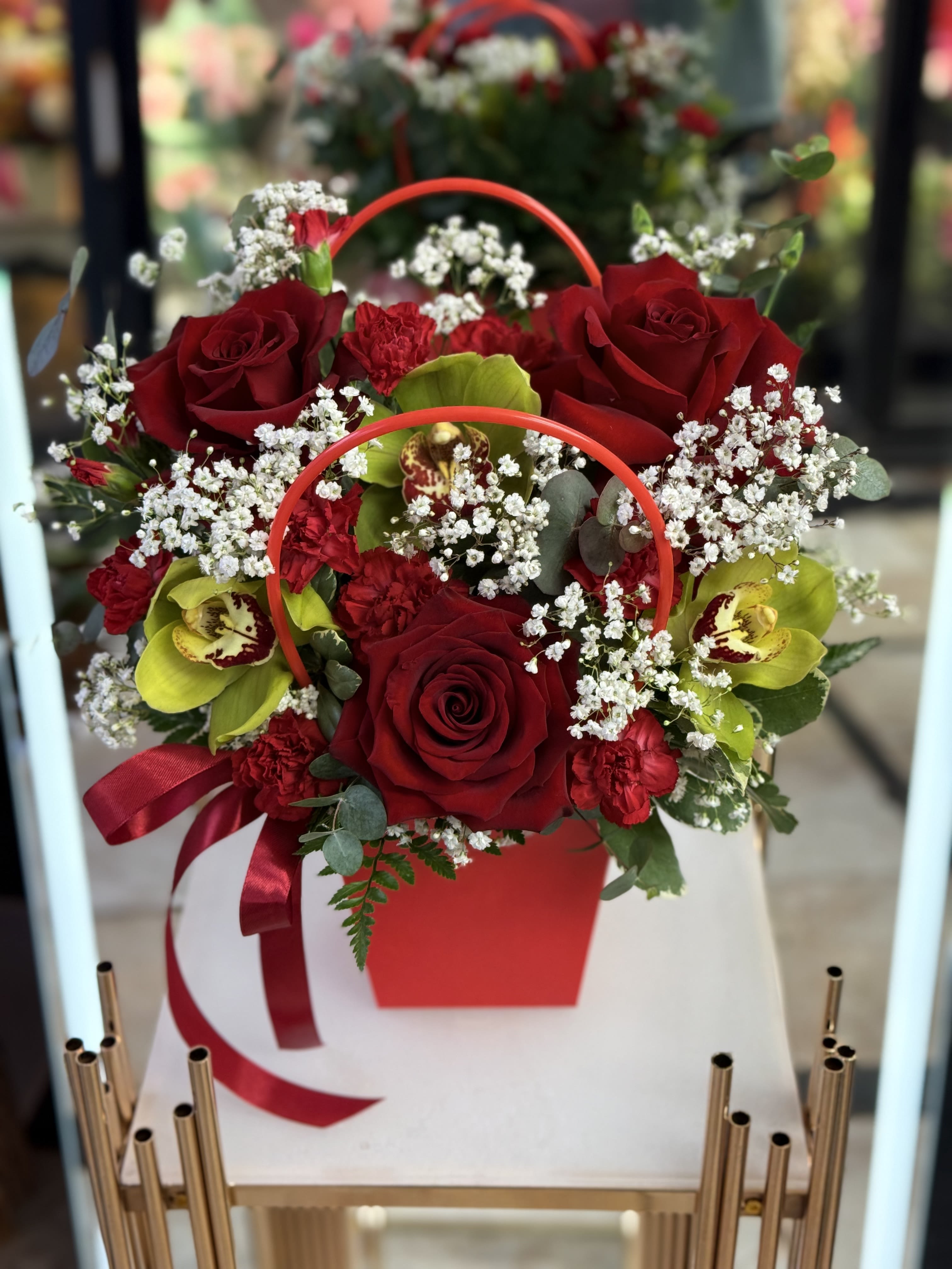 Red roses and orchids arranged in a red handled box with a ribbon