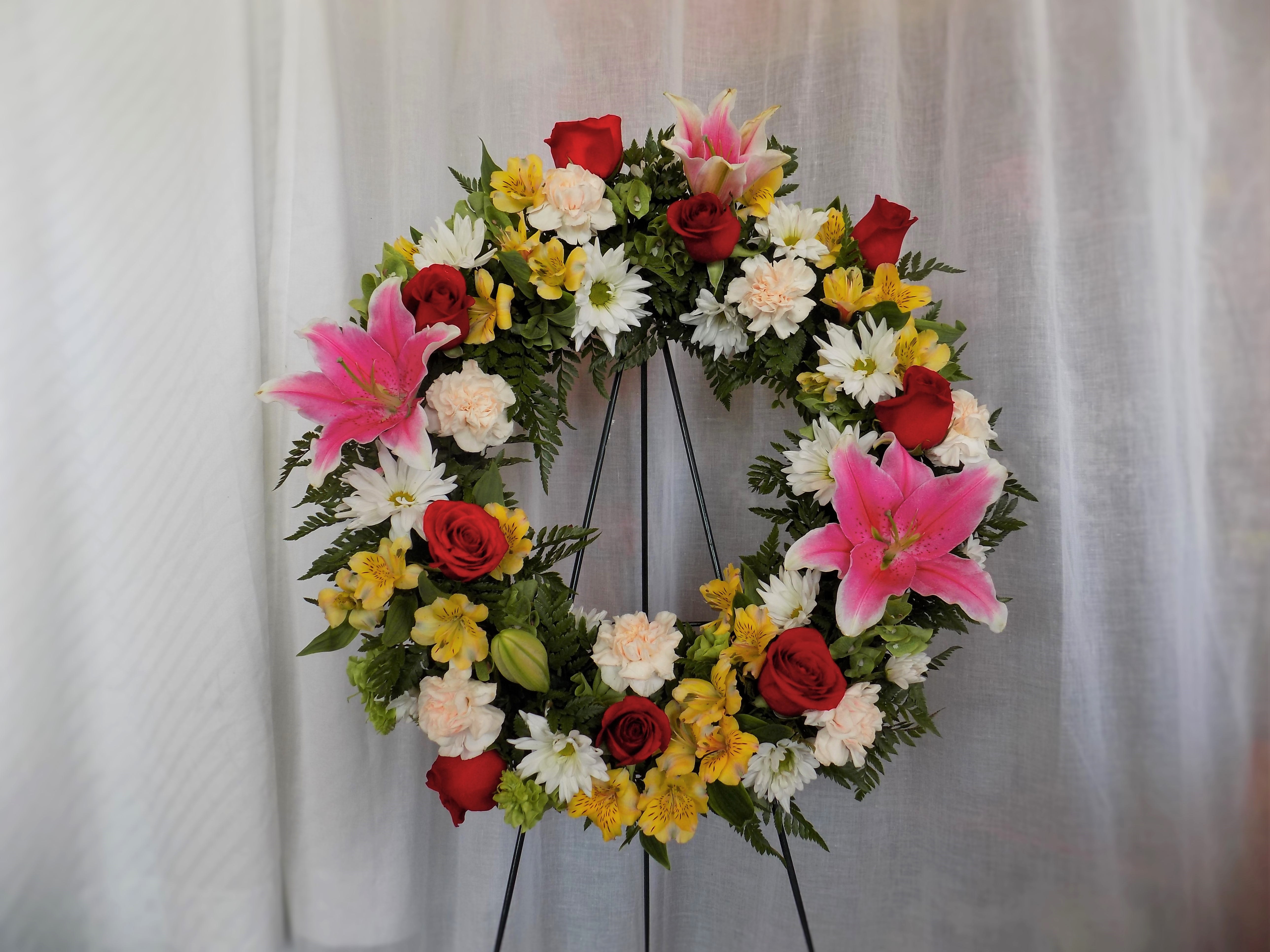 Circular floral wreath with red roses, pink lilies, and white daisies