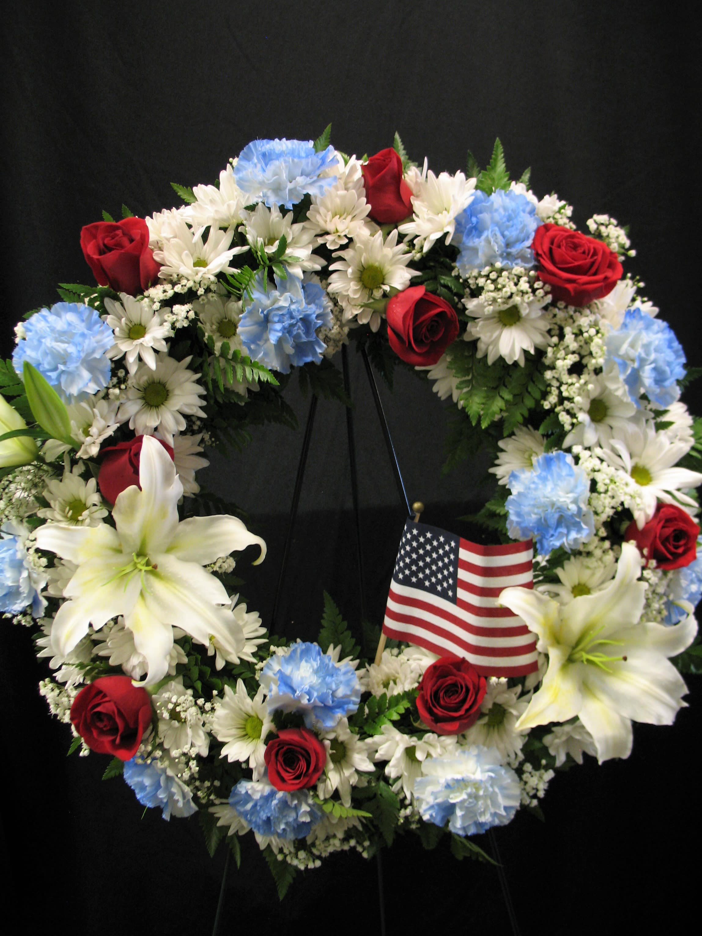 Circular wreath of red roses, white lilies, and blue carnations with a small American flag