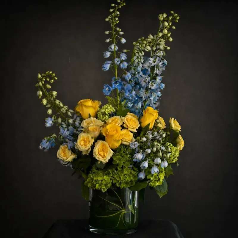 The Nurturer - A fresh and airy arrangement featuring soft blue delphinium, sunny yellow and cream roses, and lush green hydrangea with seasonal garden accents. Designed in a clear glass vase with an organic, asymmetrical silhouette for beautiful height and movement.  Each arrangement is handcrafted and may vary slightly based on seasonal availability while maintaining the overall color palette and style.