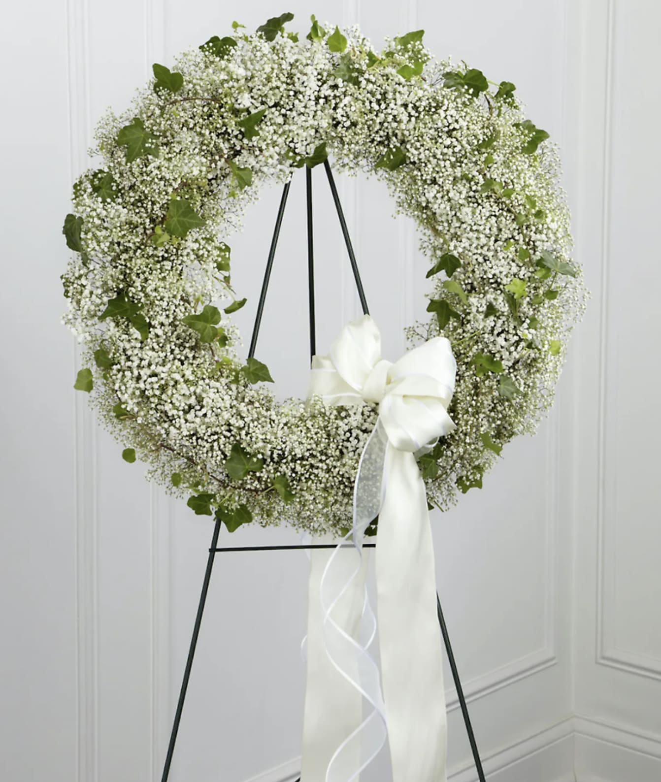 White floral wreath on a black stand with a large white ribbon bow