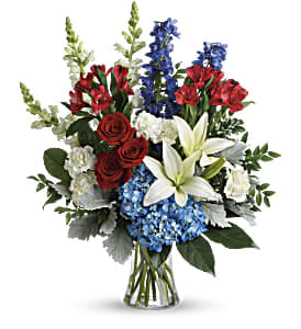 Tall red, white, and blue flower arrangement with roses, lilies, hydrangea, and delphinium in a clear glass vase